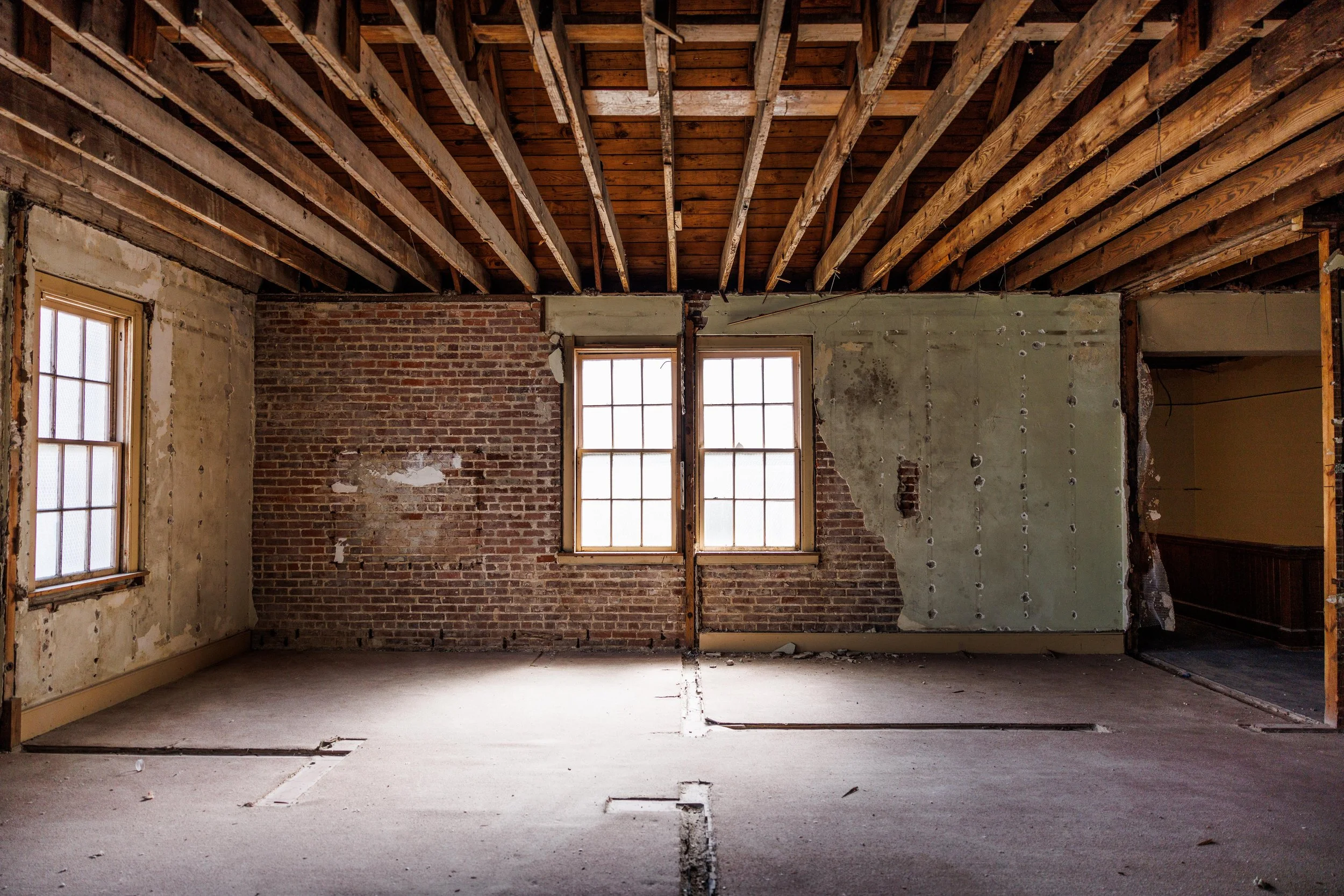 Interior of an unfinished room with exposed wooden ceiling beams, two windows, and partially removed wall exposing brick and drywall.