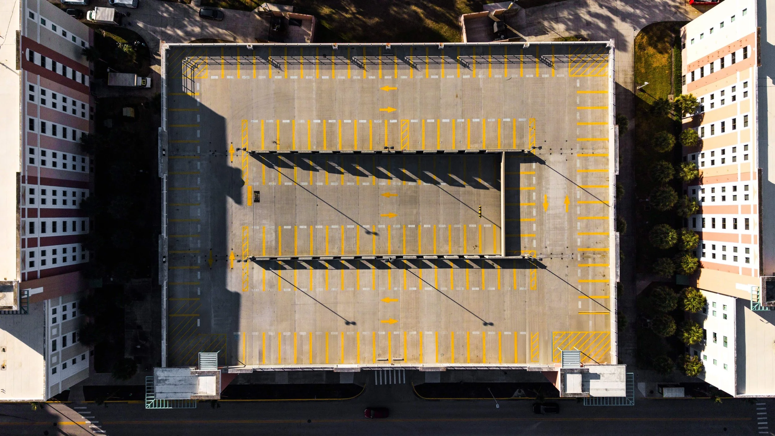 An aerial view of an empty multi-story parking garage with yellow parking lines and arrows, surrounded by residential buildings and trees.