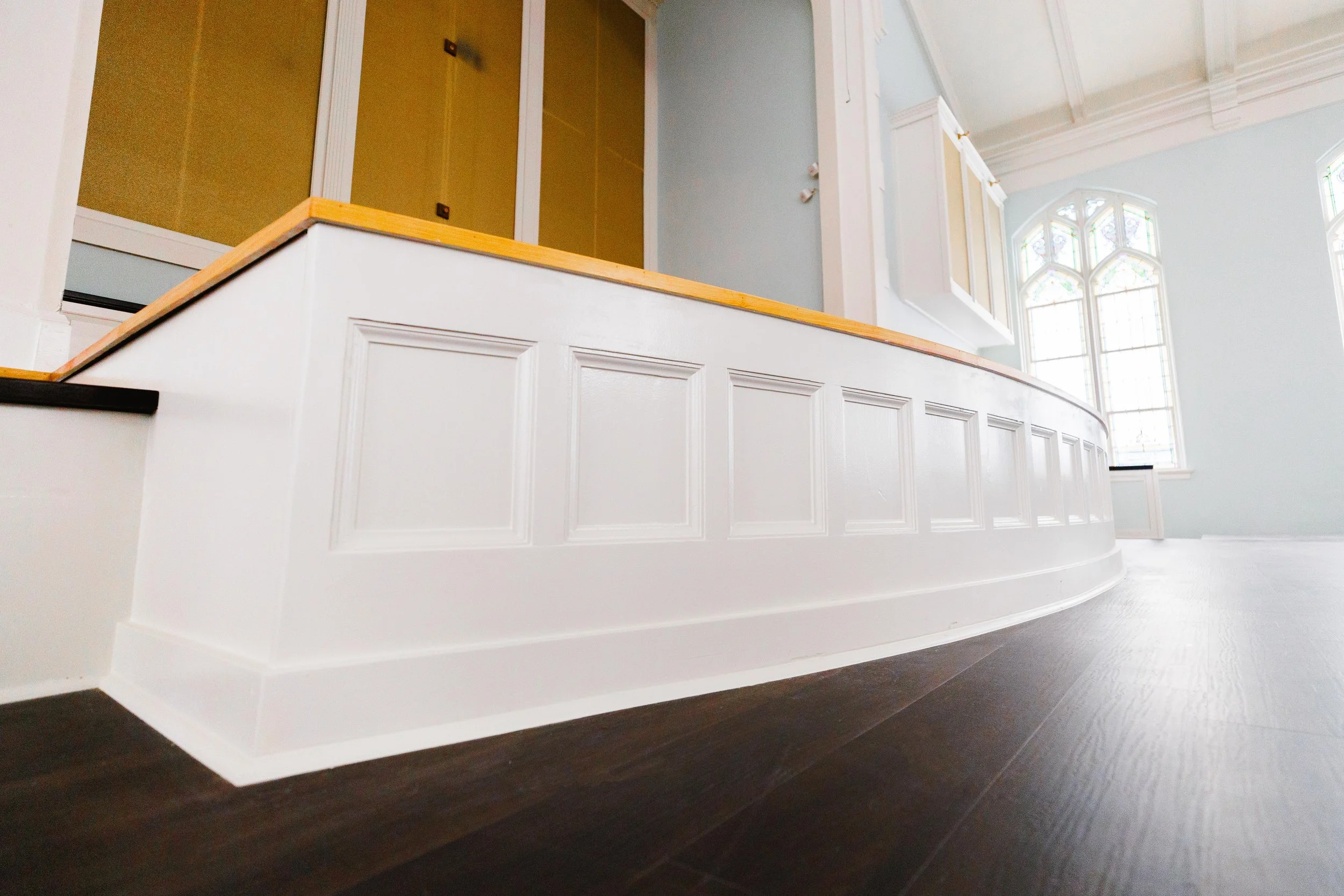 Interior view of a room with a curved white wooden panel with recessed rectangular designs, dark hardwood flooring, large stained glass windows, and a partial view of a balcony or elevated area with a yellow railing.