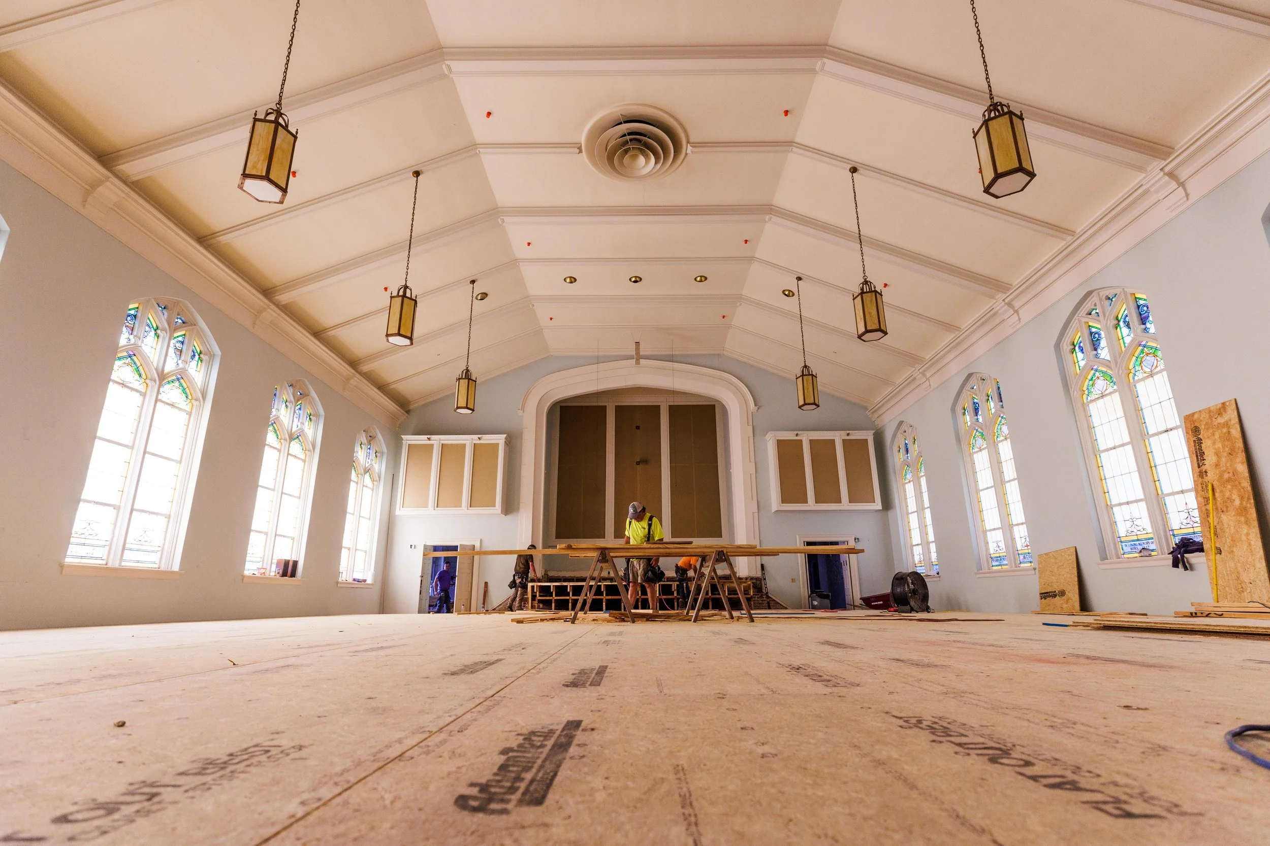 Construction workers working inside a large church or hall with stained glass windows, hanging pendant lights, and a high vaulted ceiling.