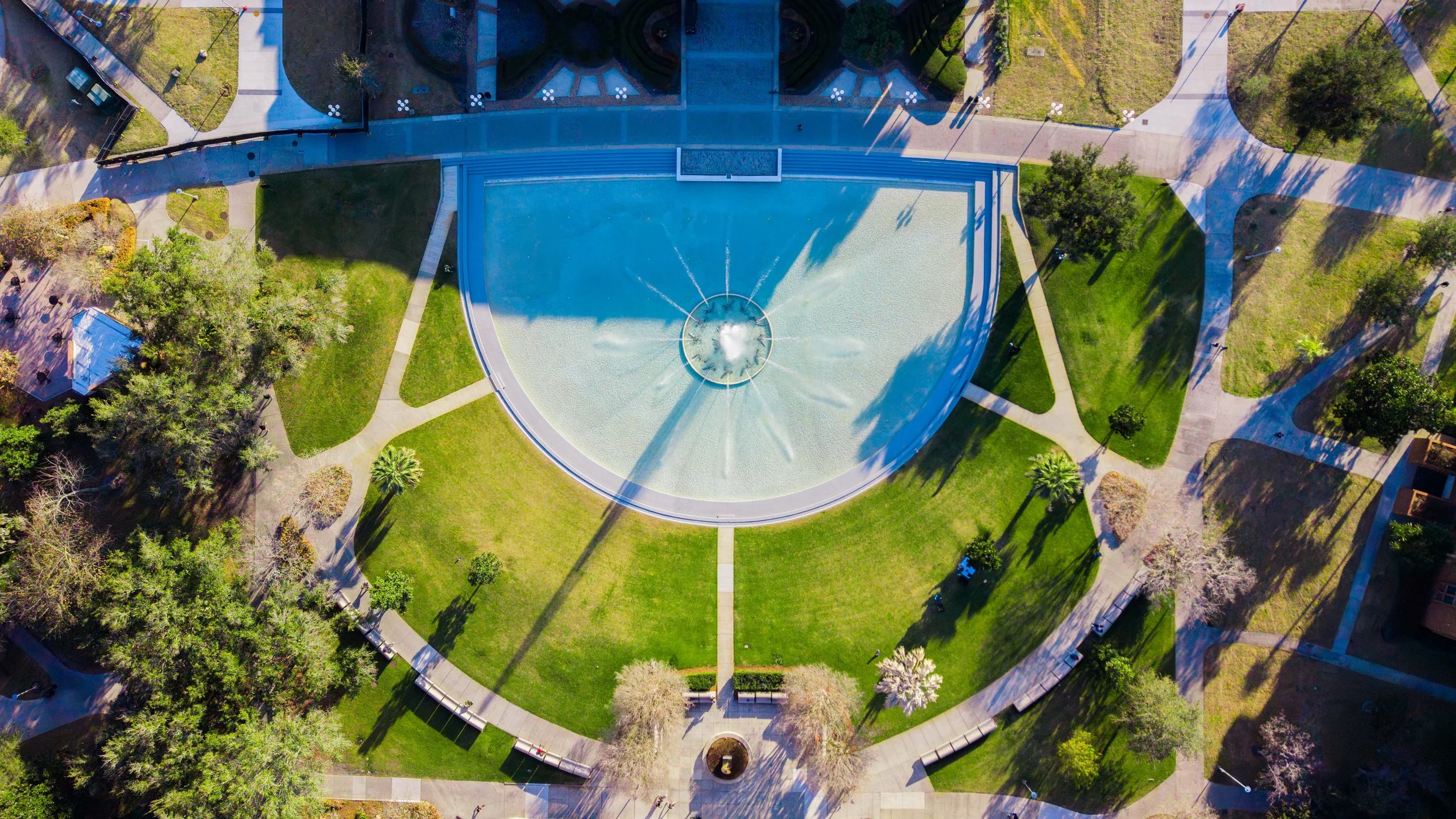 An aerial view of a park features a large, semi-circular fountain with water jets in the center, surrounded by grassy lawns, trees, walkways, benches, and a building at the top.