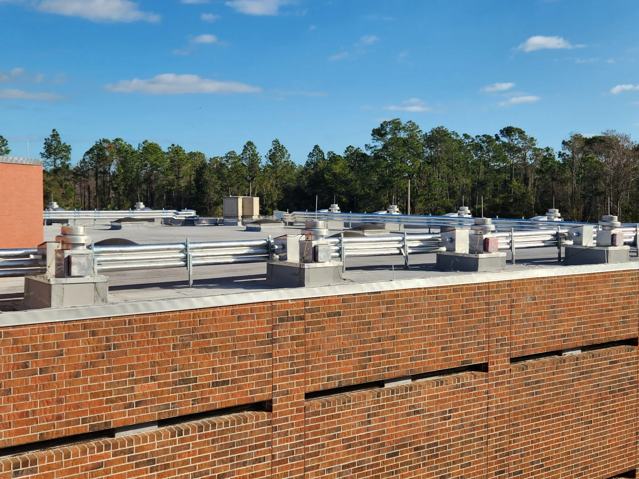 Rooftop with metal vents and ductwork on a brick building under a partly cloudy sky, with trees in the background.