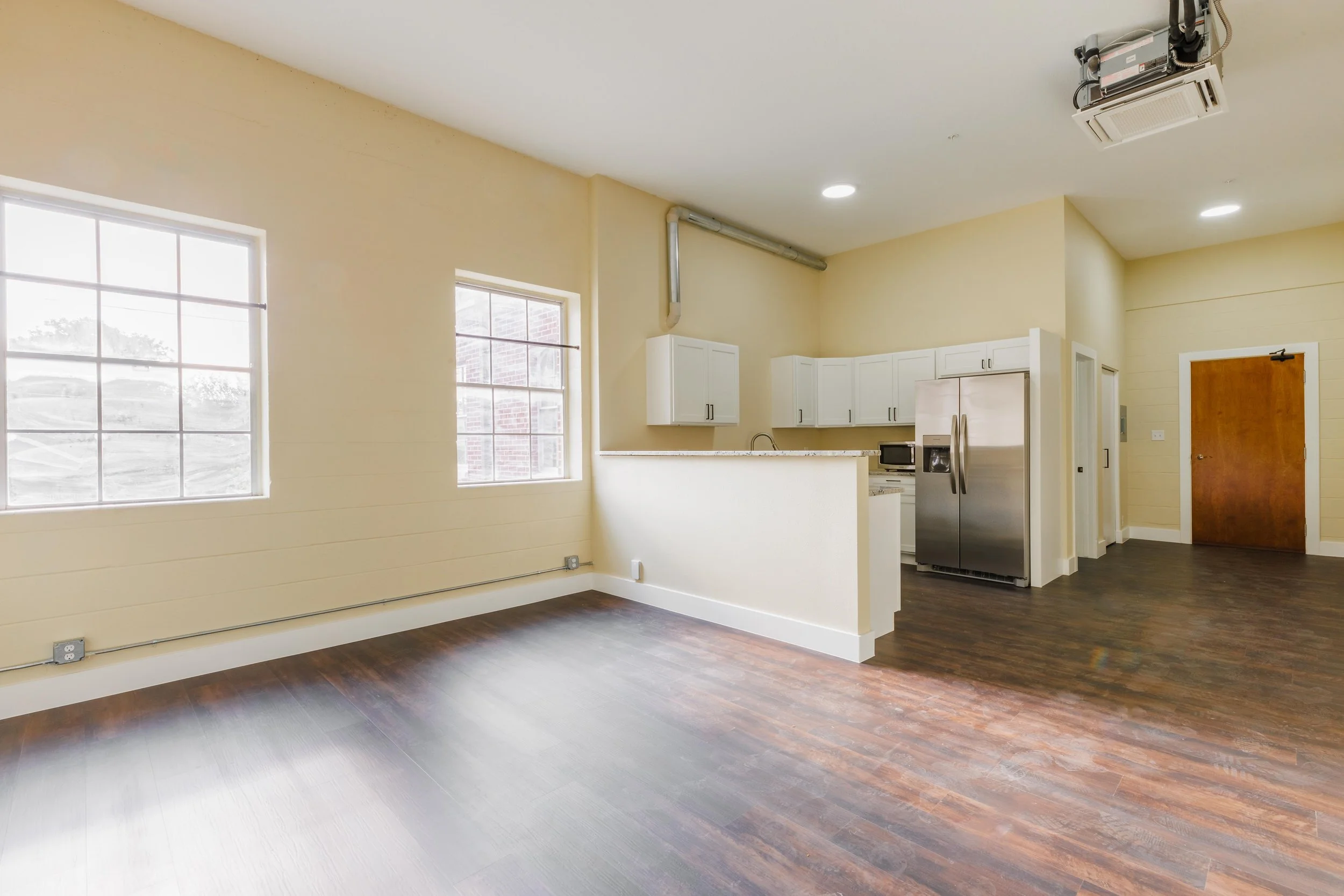Empty living room with dark hardwood floors, cream-colored walls, and two large windows. The kitchen area has white cabinets, a stainless steel refrigerator, and a microwave, with a small breakfast bar.