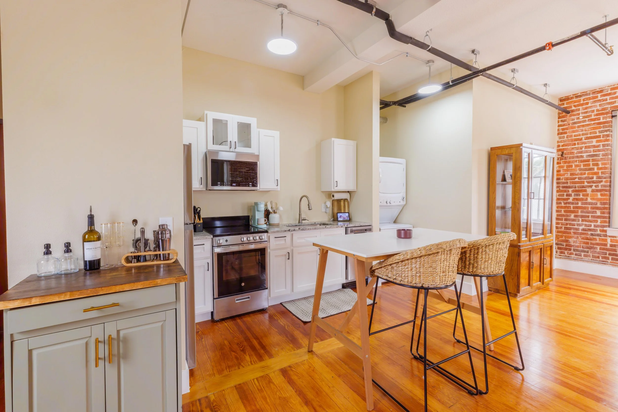 Open-concept kitchen with white cabinets, stainless steel appliances, a small dining island with wicker chairs, hardwood floors, exposed brick wall, and hanging ceiling lights.