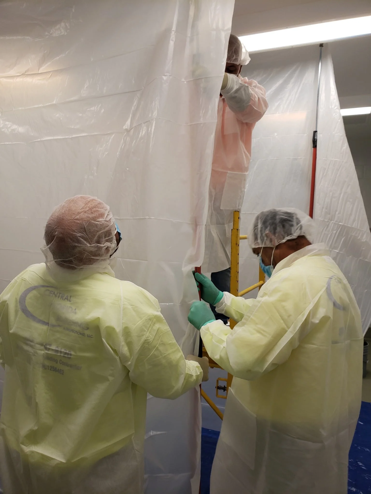 Three healthcare workers in yellow gowns, masks, gloves, and hair covers working inside a medical setting with white plastic walls, possibly during a procedure or assembly.