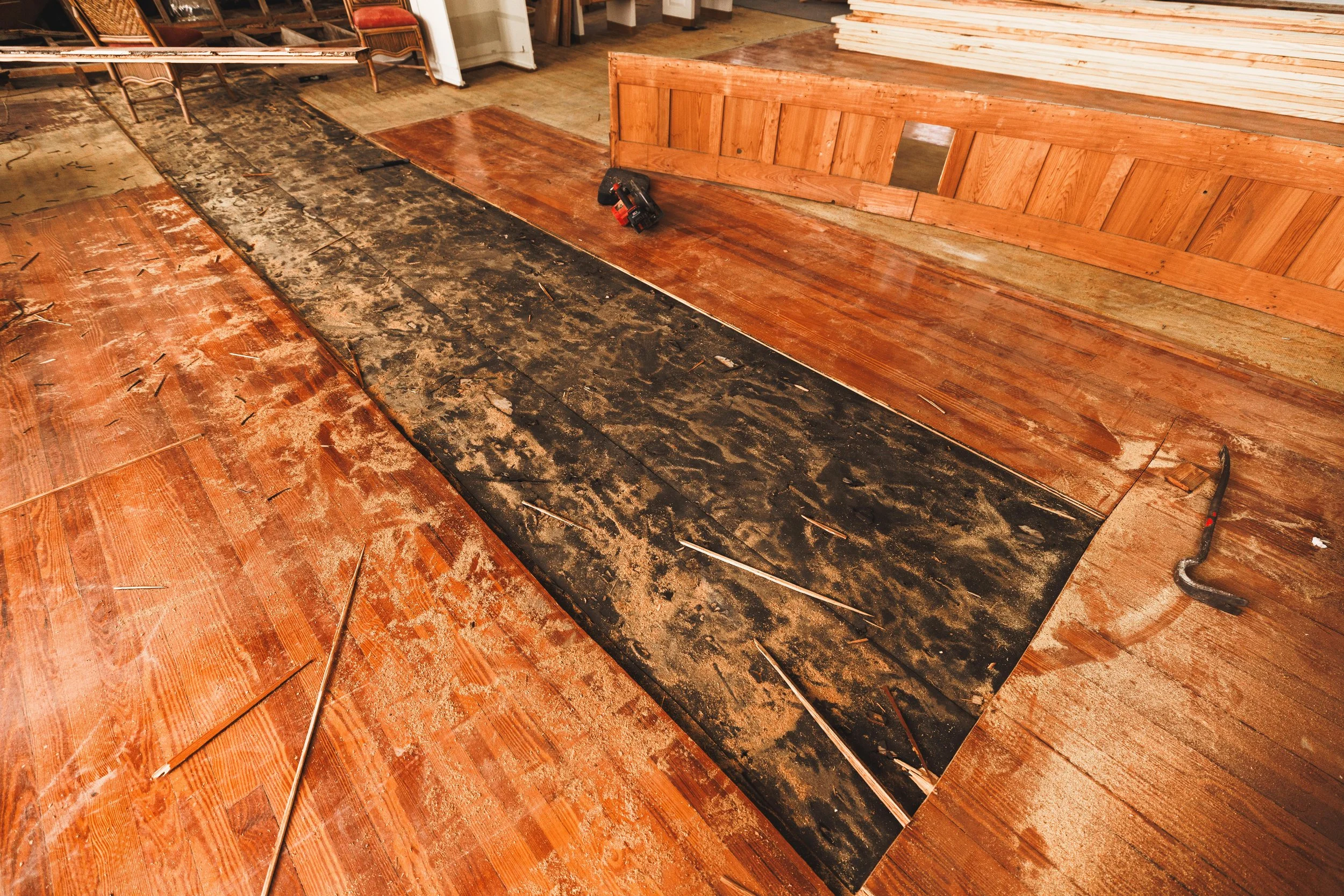 A woodworking workspace showing a partially sanded hardwood floor, with a black protective mat in the middle and tools nearby.