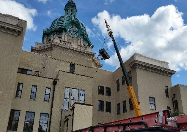 Construction crane lifting a firefighter's basket near a historic building with a clock tower against a blue sky with clouds.