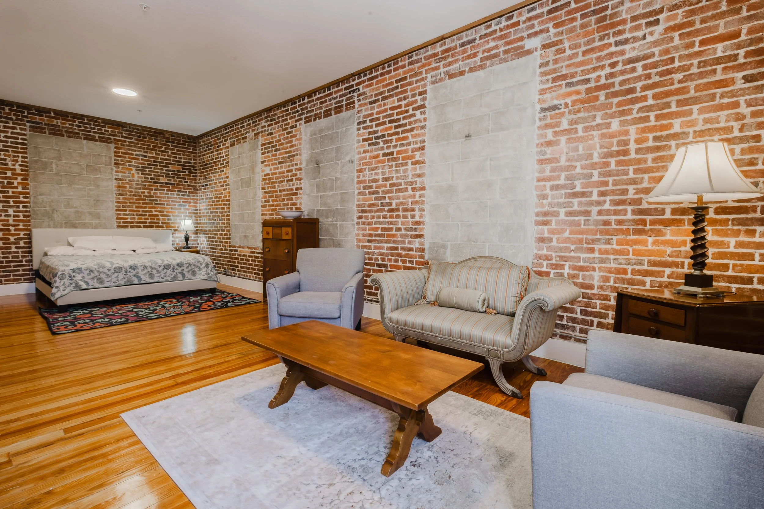 A spacious living area with brick and stone accent walls, a bed in the corner, and vintage-style furniture including armchairs, a sofa, a wooden coffee table, and side tables with lamps.