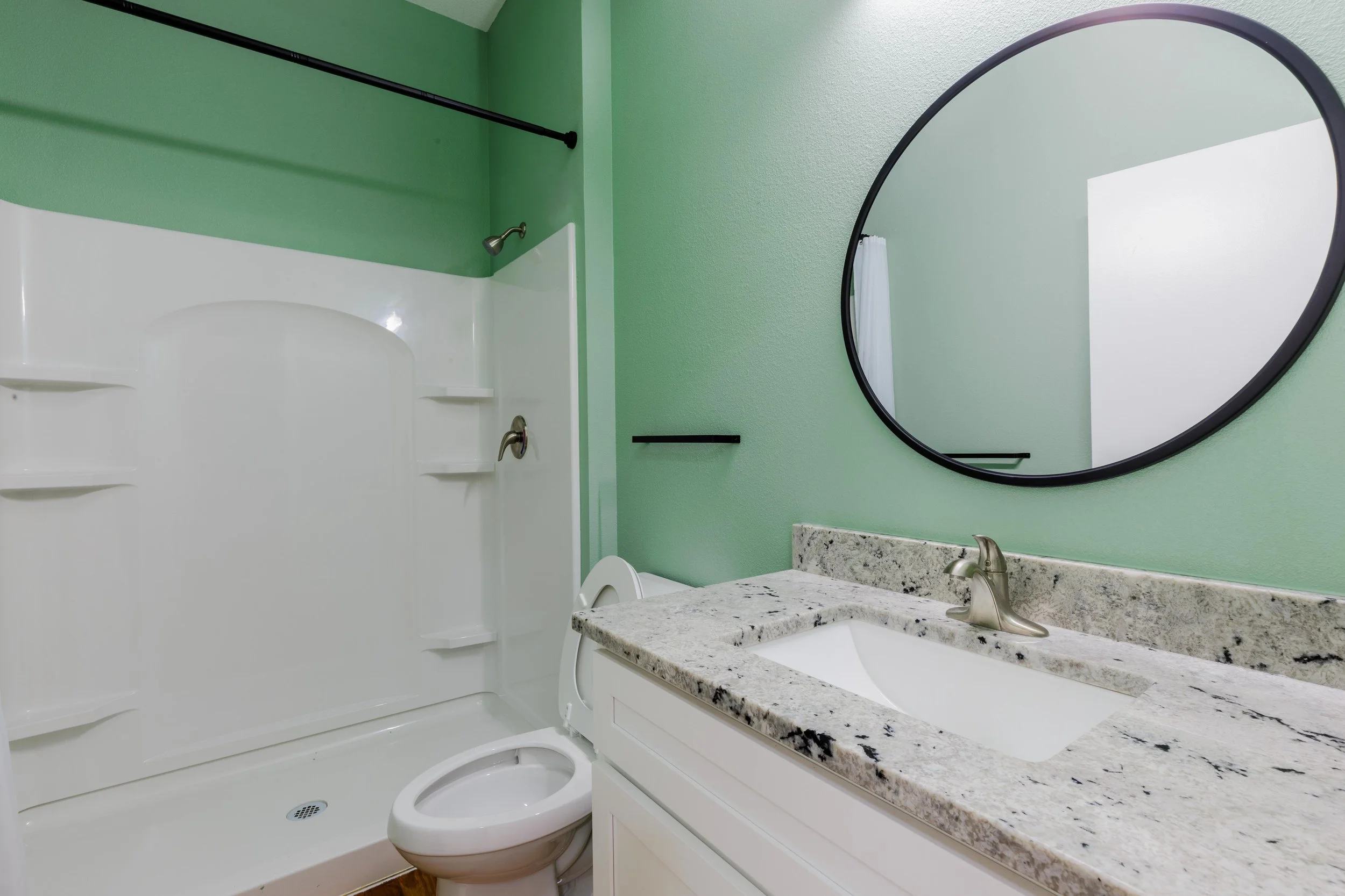 Bathroom with a shower area, toilet, granite countertop with a sink, and a large round mirror on a green wall