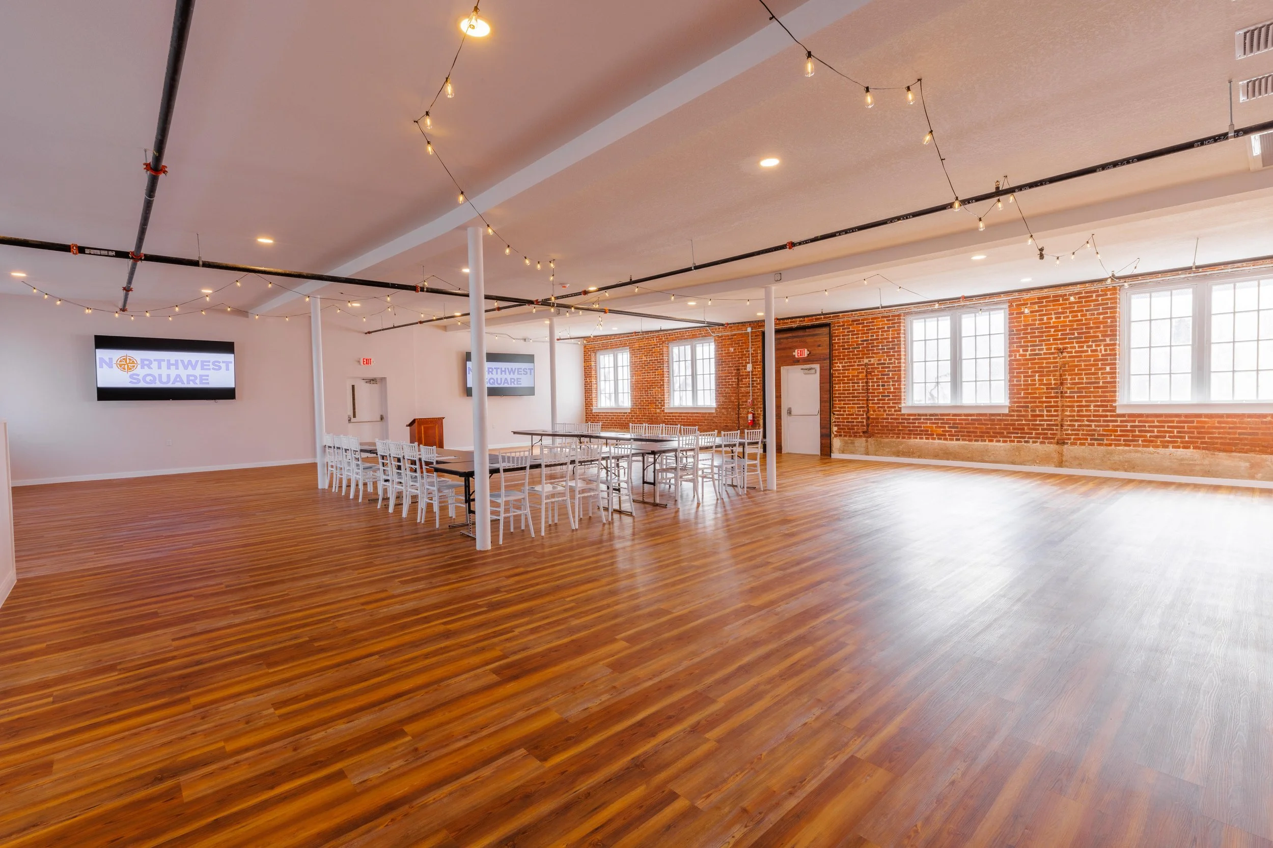 An empty event space with wooden floors, white walls, exposed brick wall, multiple windows, string lights hanging from the ceiling, and television screens displaying 'Northwest Square'. There are white chairs and tables set up in the center of the ro