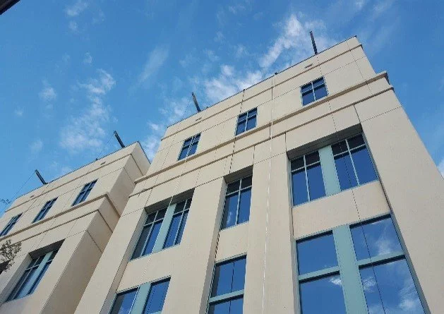 Looking up at a modern multi-story beige building with large blue-tinted windows against a partly cloudy blue sky.