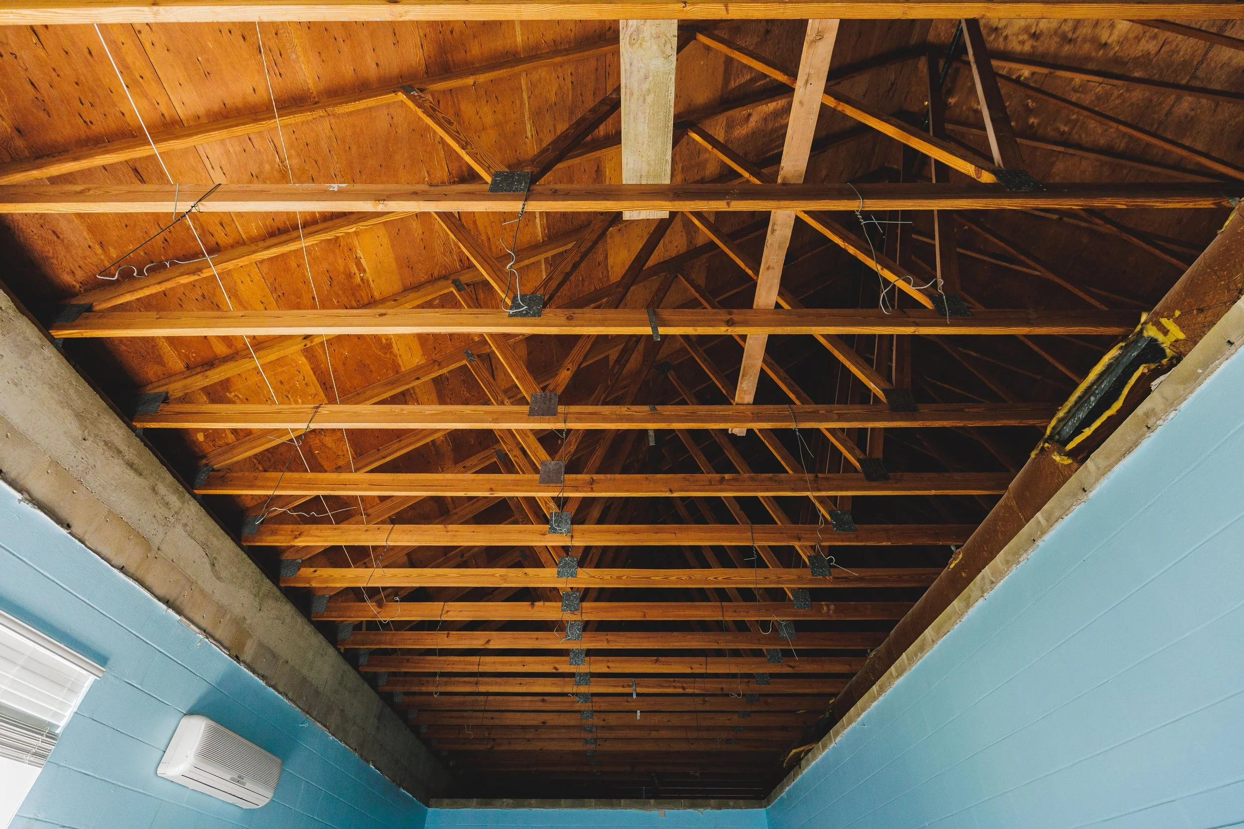 View of a building's exposed wooden ceiling framework with electrical wires, blue walls, and an HVAC unit in the lower left corner.