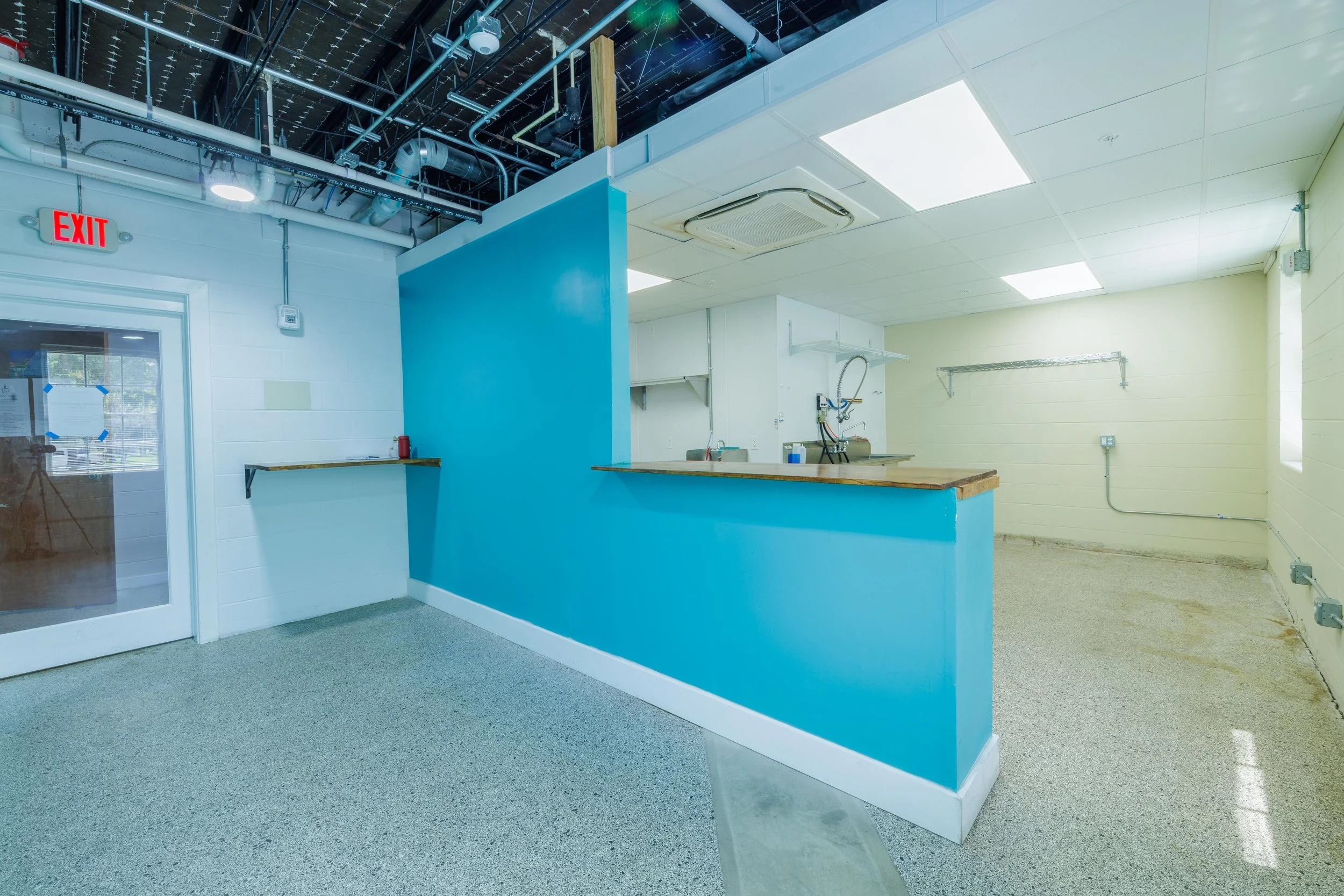 Interior of a commercial or industrial space with a blue partition wall, a counter with a wooden top, a sink, shelves, and a ceiling with exposed ductwork and lighting panels. An exit door is visible on the left.