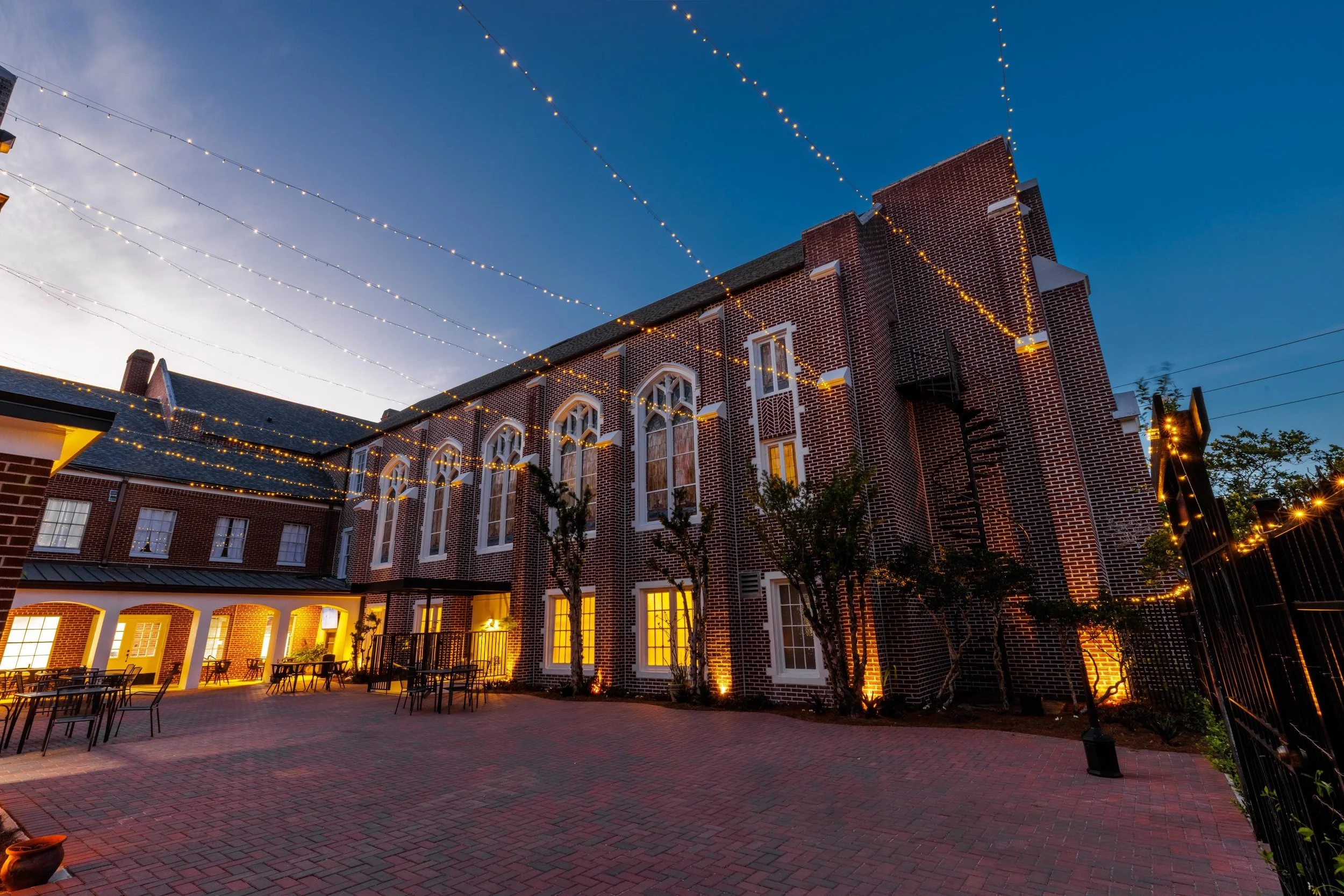 Outdoor courtyard outside a brick building decorated with string lights at dusk, with tables and trees.