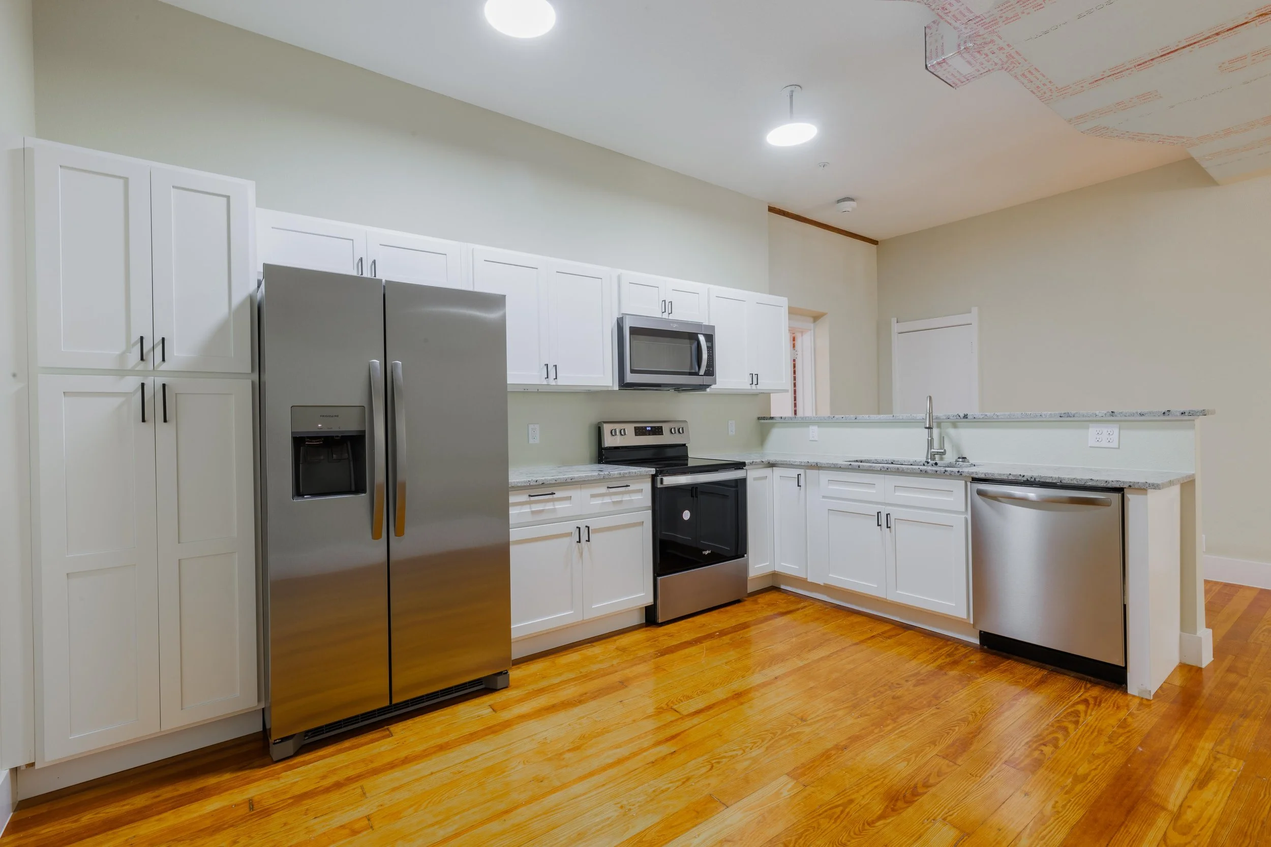 Empty modern kitchen with white cabinets, stainless steel appliances, granite countertops, and hardwood floors.