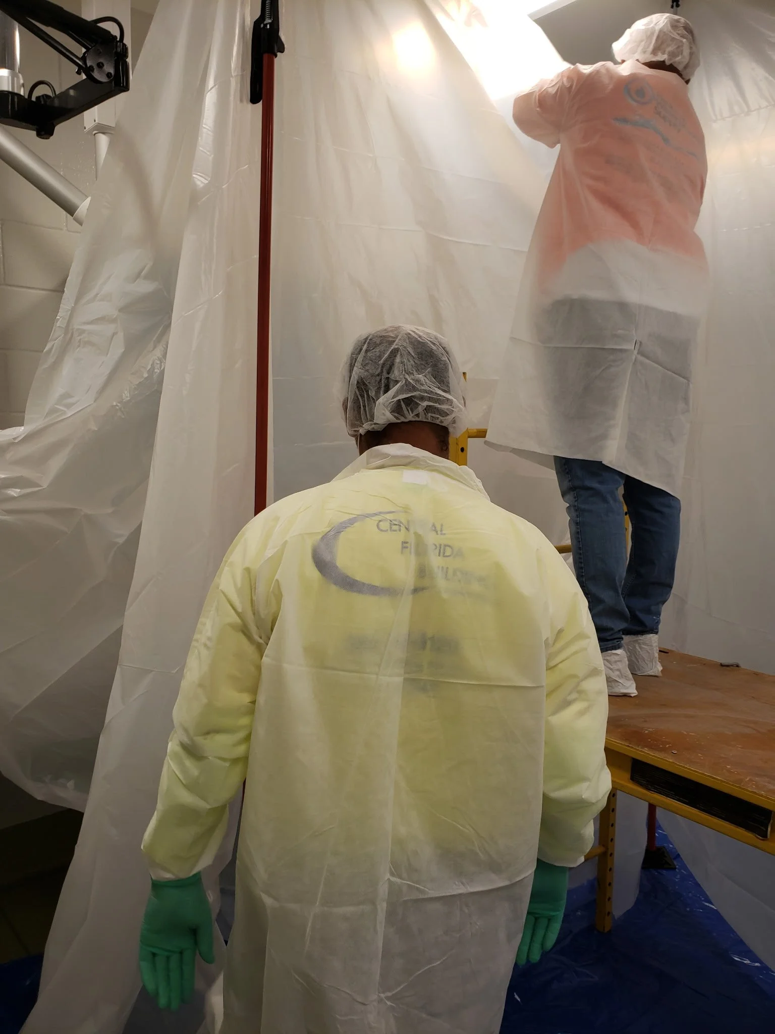 Medical workers in protective gear working inside a sterile environment, with plastic sheeting and equipment visible.