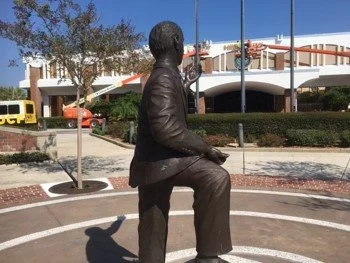 Bronze statue of a man in a suit with one foot raised, standing on a circular base outdoors with trees, buildings, and cars in the background.