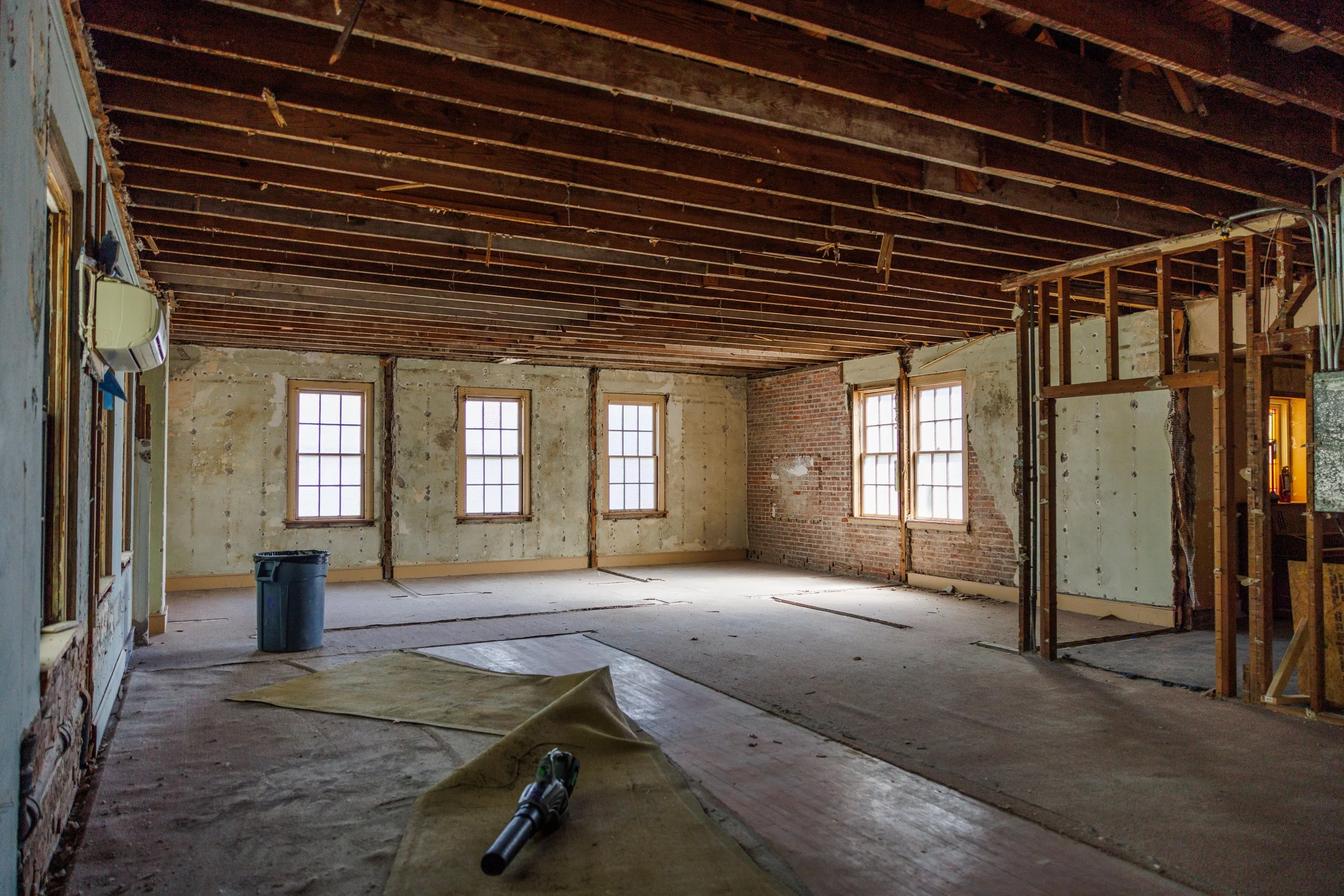 Interior of a room under renovation with exposed wooden ceiling beams, three windows, partially removed drywall revealing brick wall, construction tools, and debris on the floor.