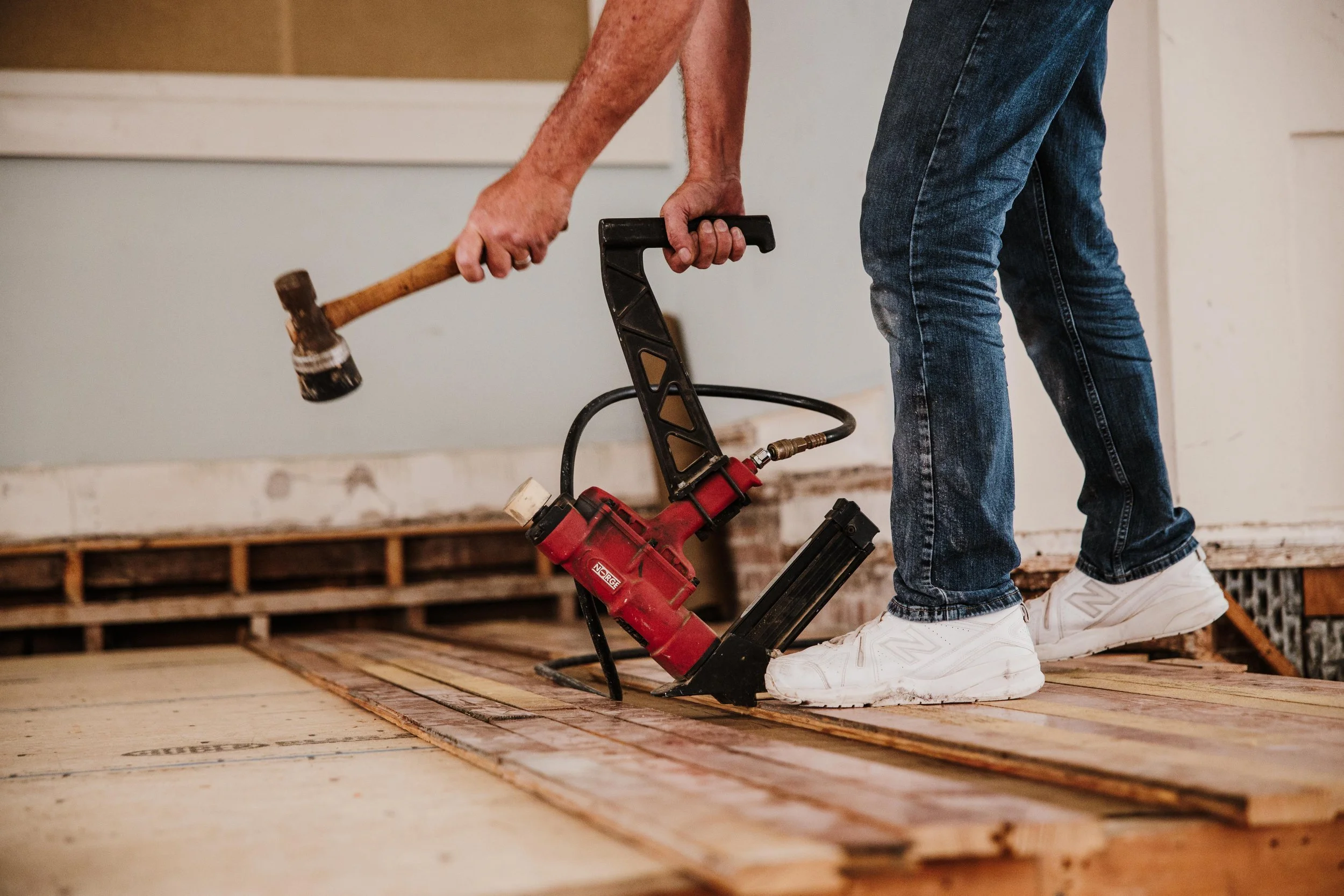 Person wearing blue jeans and white sneakers using a hammer to install wooden flooring while standing on the new floor.