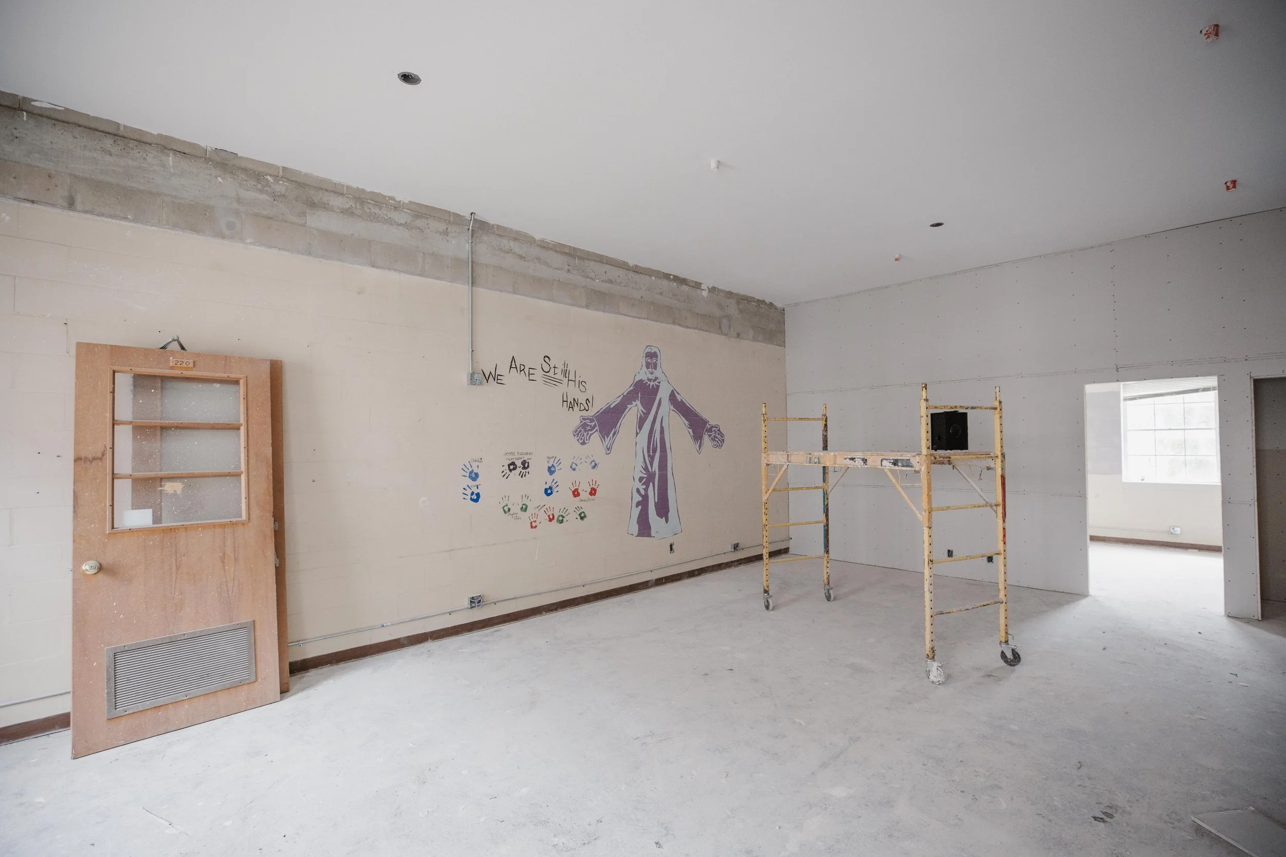 Interior of a room under construction with unfinished walls and ceiling, a wooden door leaning against the wall, a scaffold in the center, a window in the background, and colorful wall art with a person and the text "We Still this Hands!"