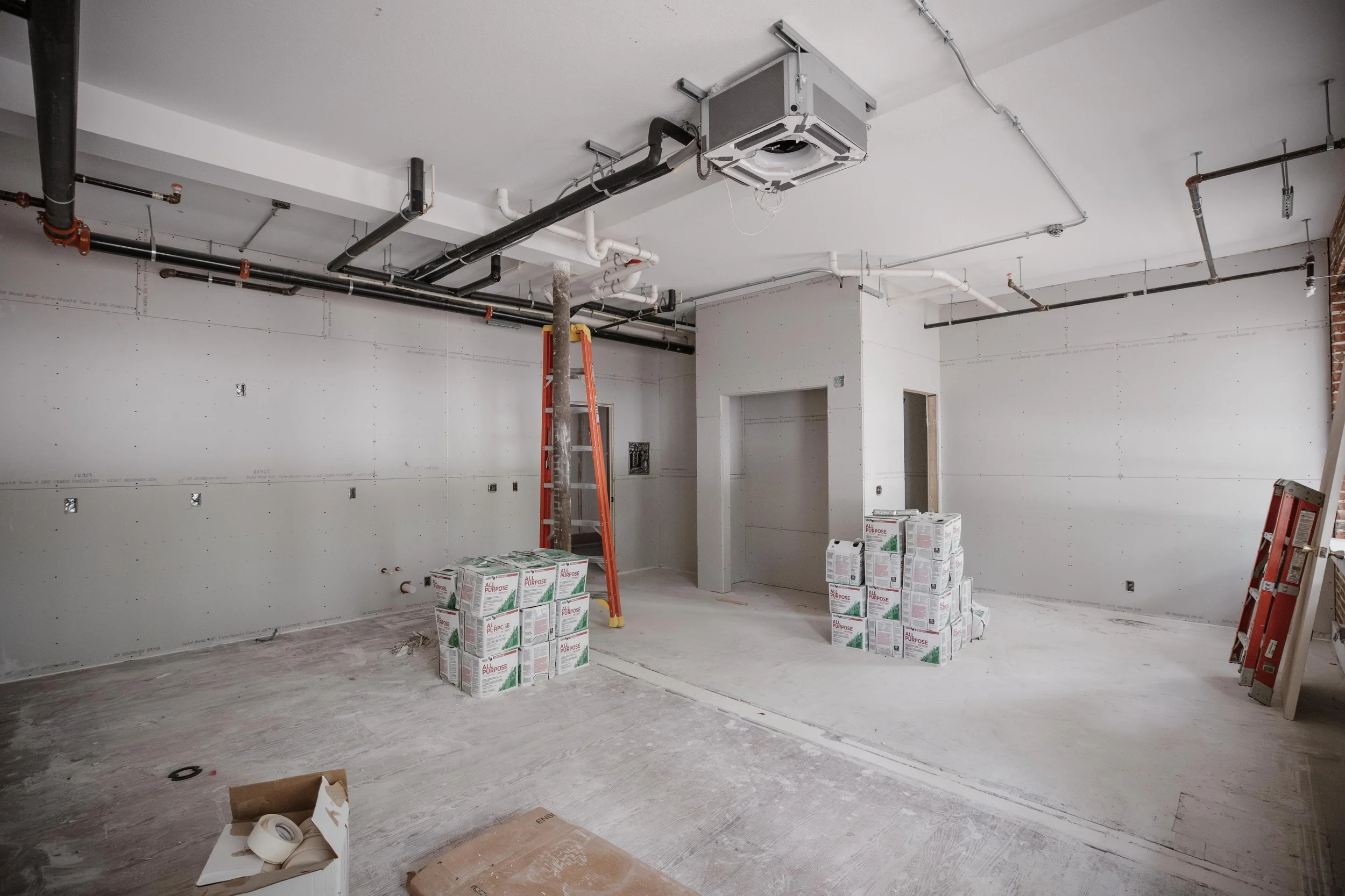 An unfinished interior room under construction with bare walls, exposed pipes, a ladder, and stacked boxes of construction supplies.