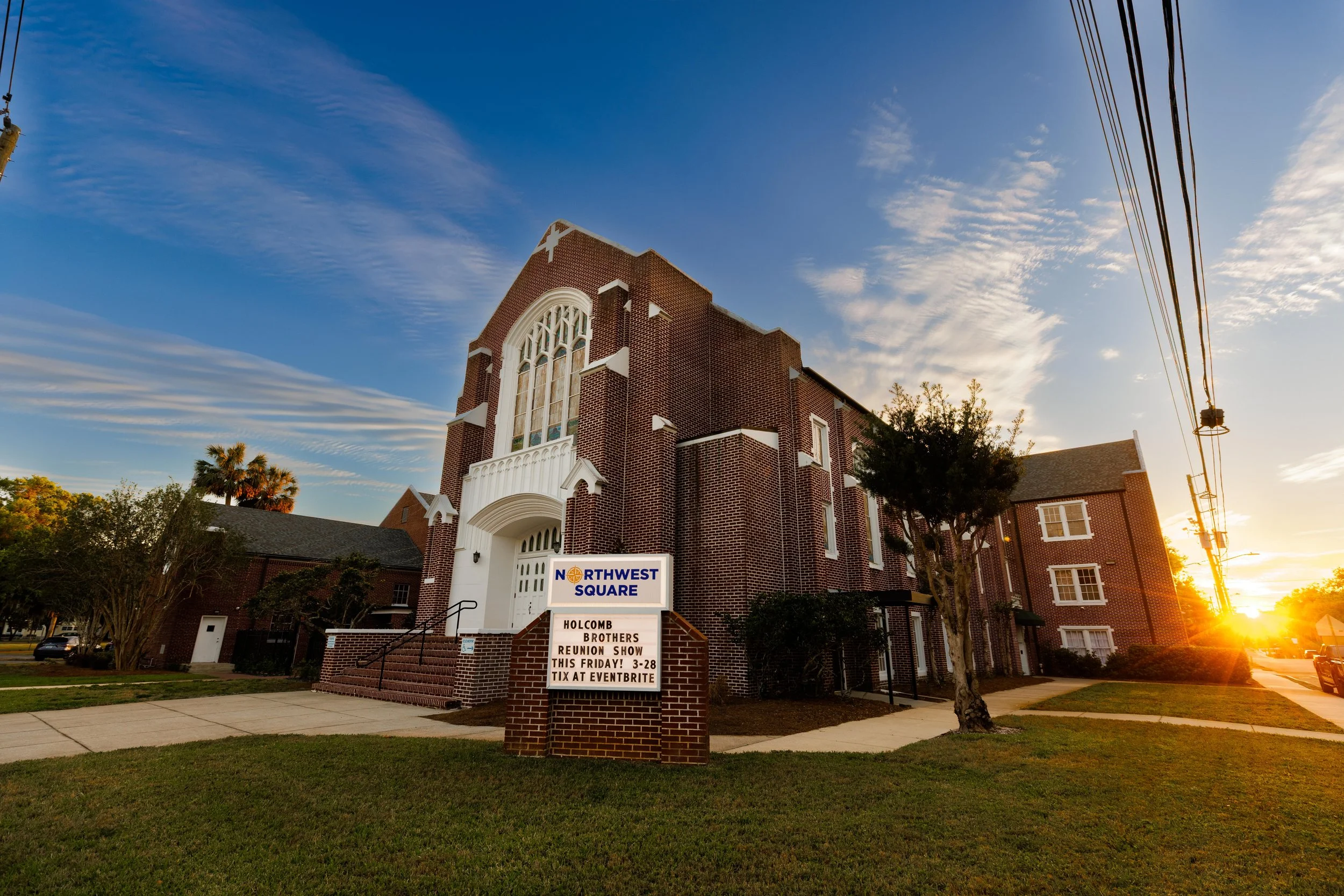 A brick church building with large arched windows and a sign on the lawn. The sign reads 'Northwest Square' and advertises a reunion show at the church.
