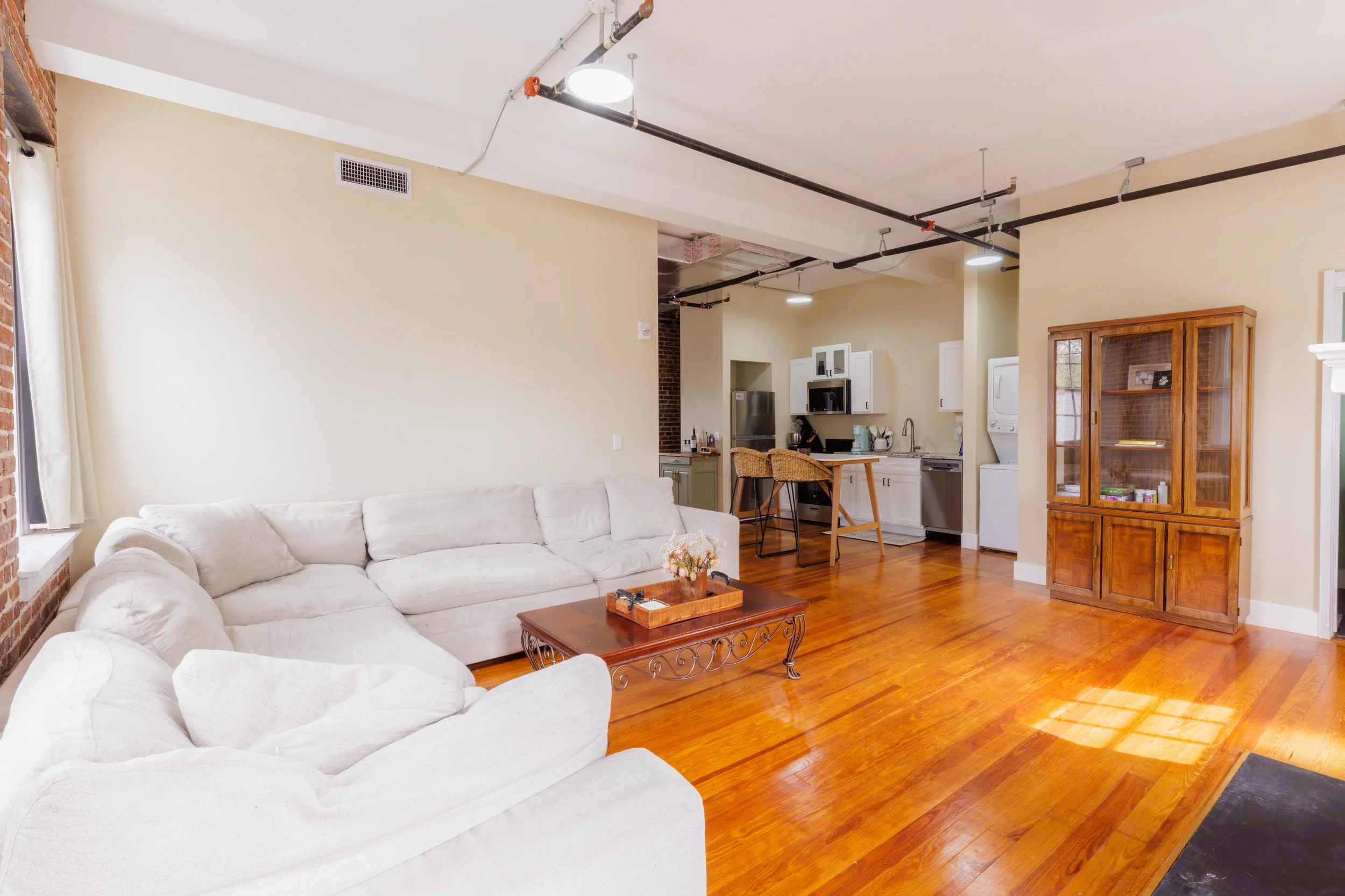 Living room with a white sectional sofa, wooden coffee table with decorative tray and flowers, and a wooden cabinet. The background features a kitchen with white cabinets, a stainless steel fridge, and a built-in microwave. There are wooden bar stool