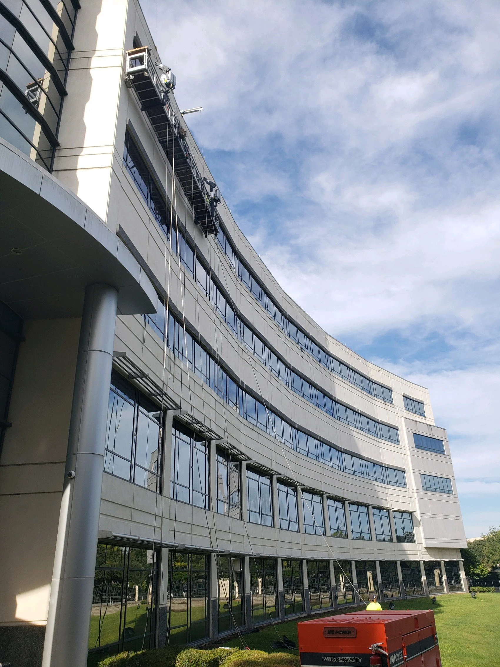 Construction workers are cleaning windows on a modern, curved building with a blue sky and clouds in the background.