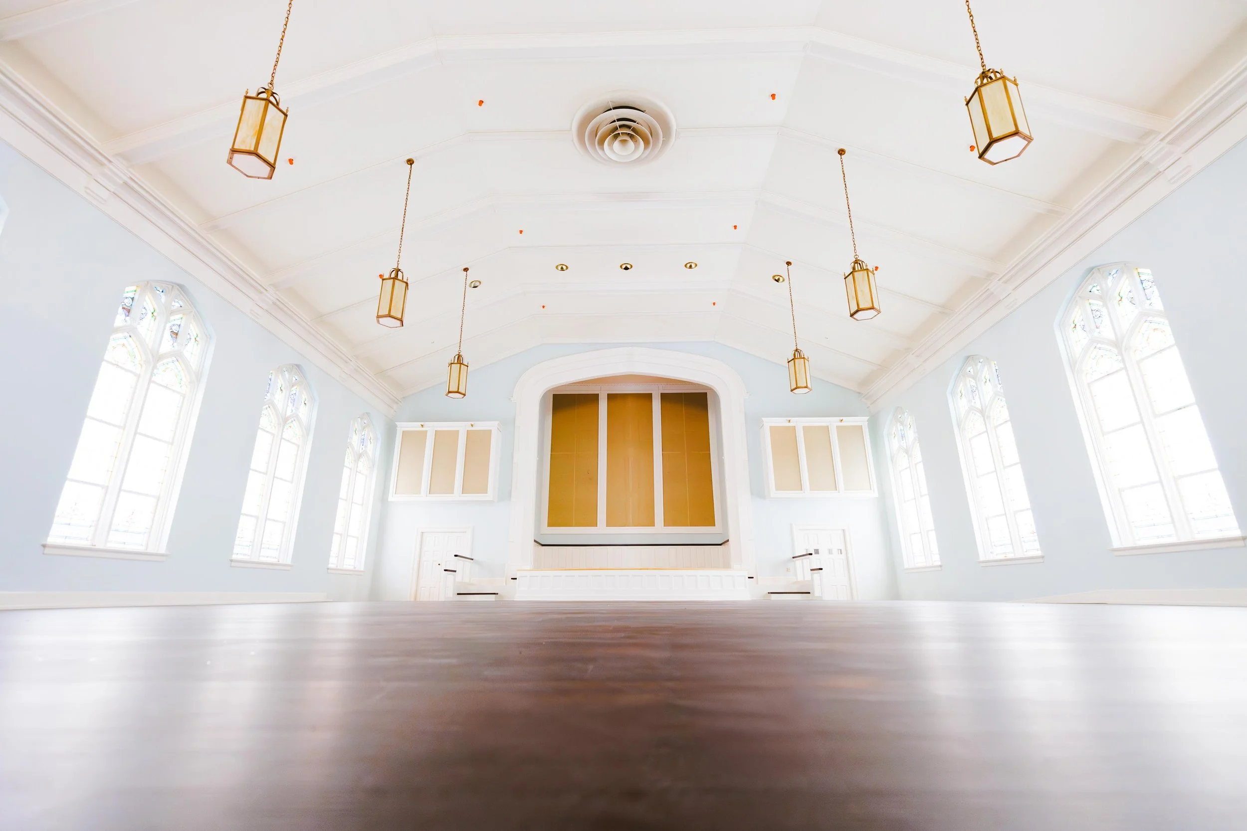 View of an empty, spacious church with high vaulted ceiling, large windows with stained glass, hanging lantern lights, and a wooden floor.
