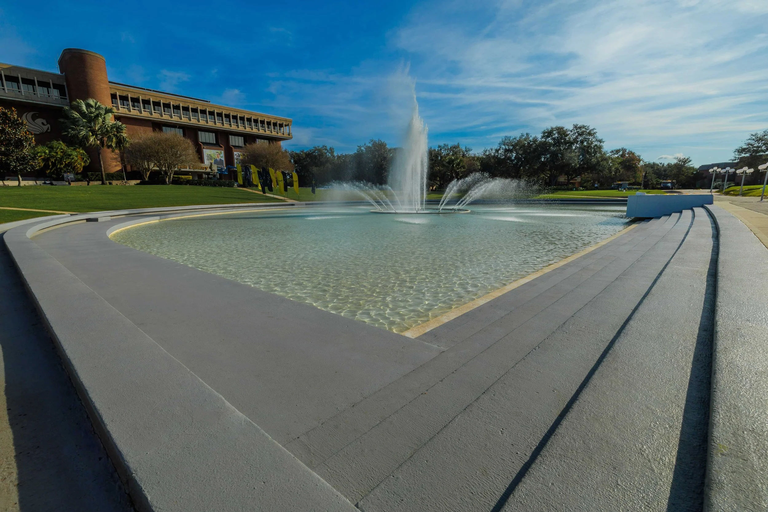 A large decorative fountain with a central jet spouting water, encircled by smaller jets, set in a wide shallow pool with clear water. The fountain is outdoors on a sunny day with a blue sky and some clouds, surrounded by green grass, trees, and a bu
