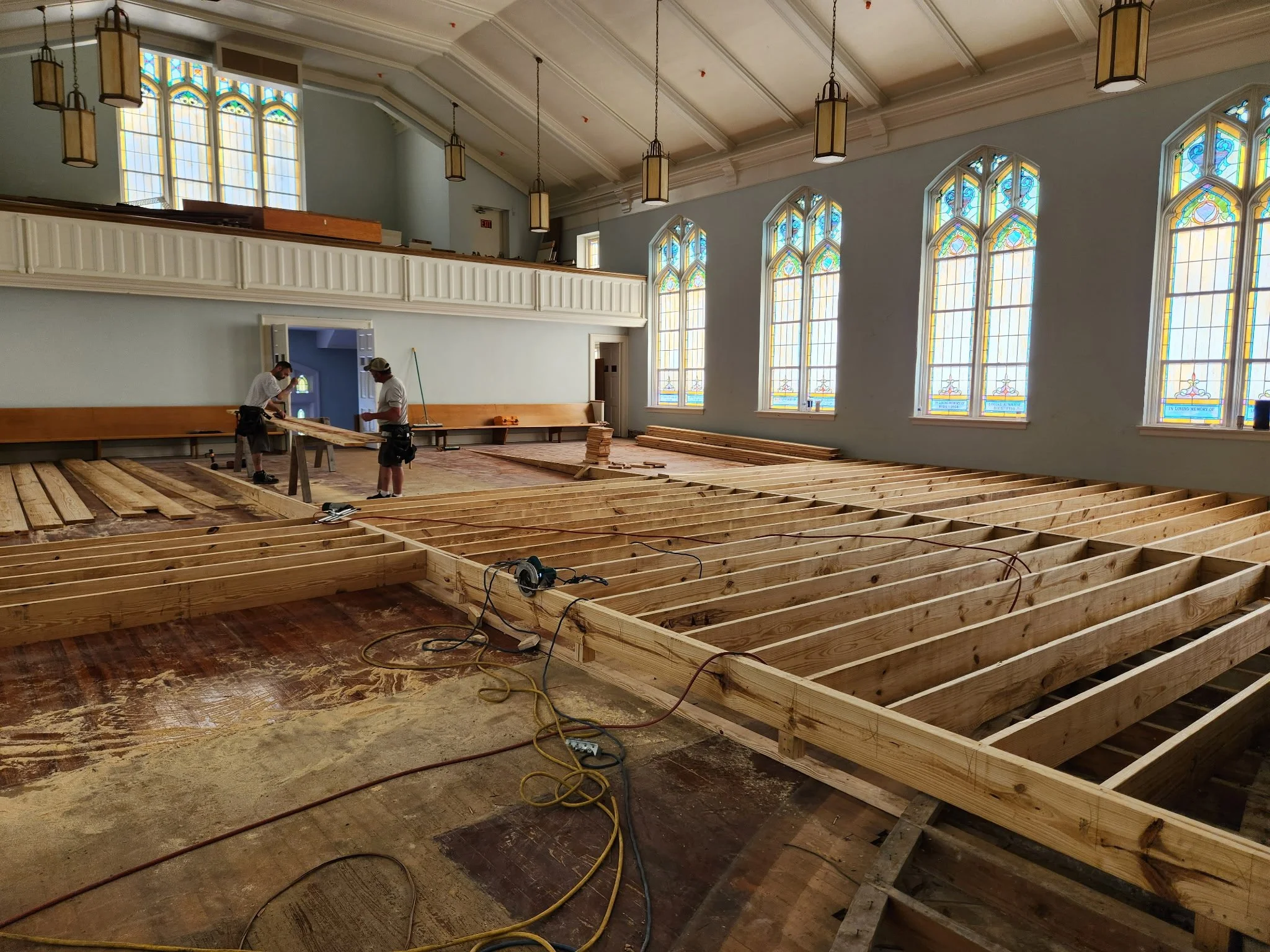 Interior of a church under renovation with exposed wooden floor framing and two workers installing the new floor. Stained glass windows are visible on the right side of the room, and electrical cords and tools are scattered across the floor.