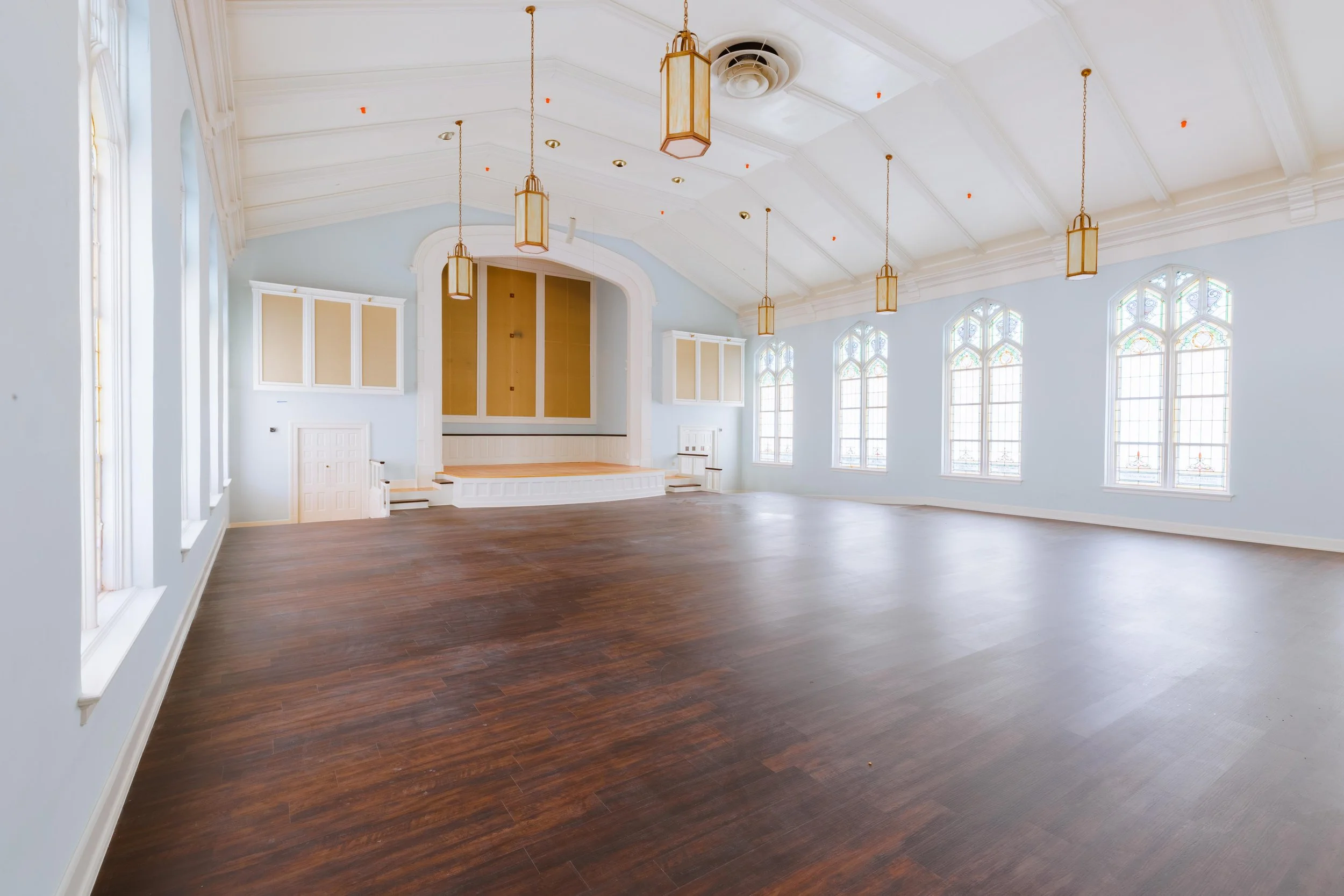 Empty, spacious room with high vaulted ceiling, large stained glass windows, wooden floor, hanging pendant lights, and a small stage at the front.