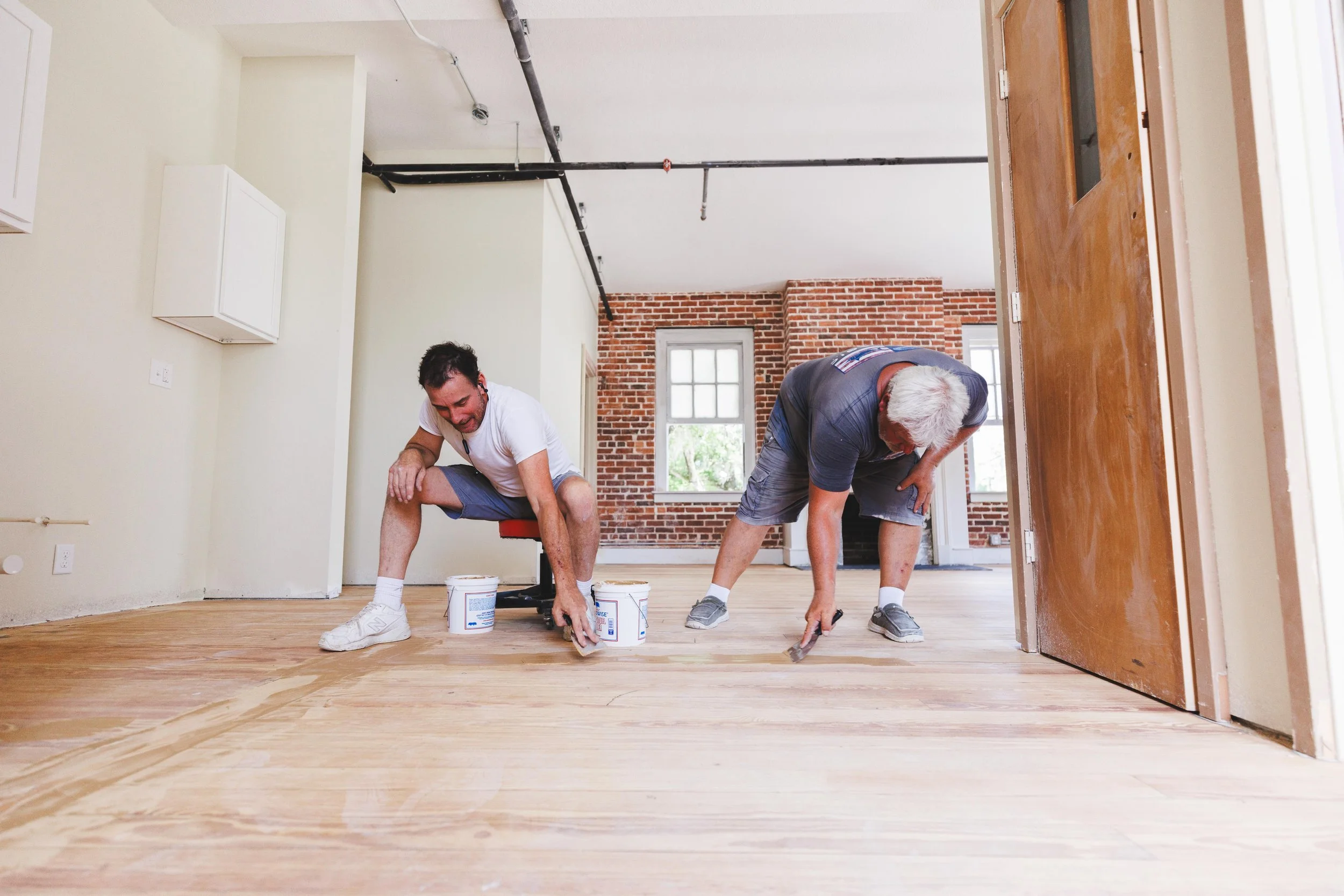 Two men working on refinishing a wooden floor inside a house, with paint cans and brushes nearby.