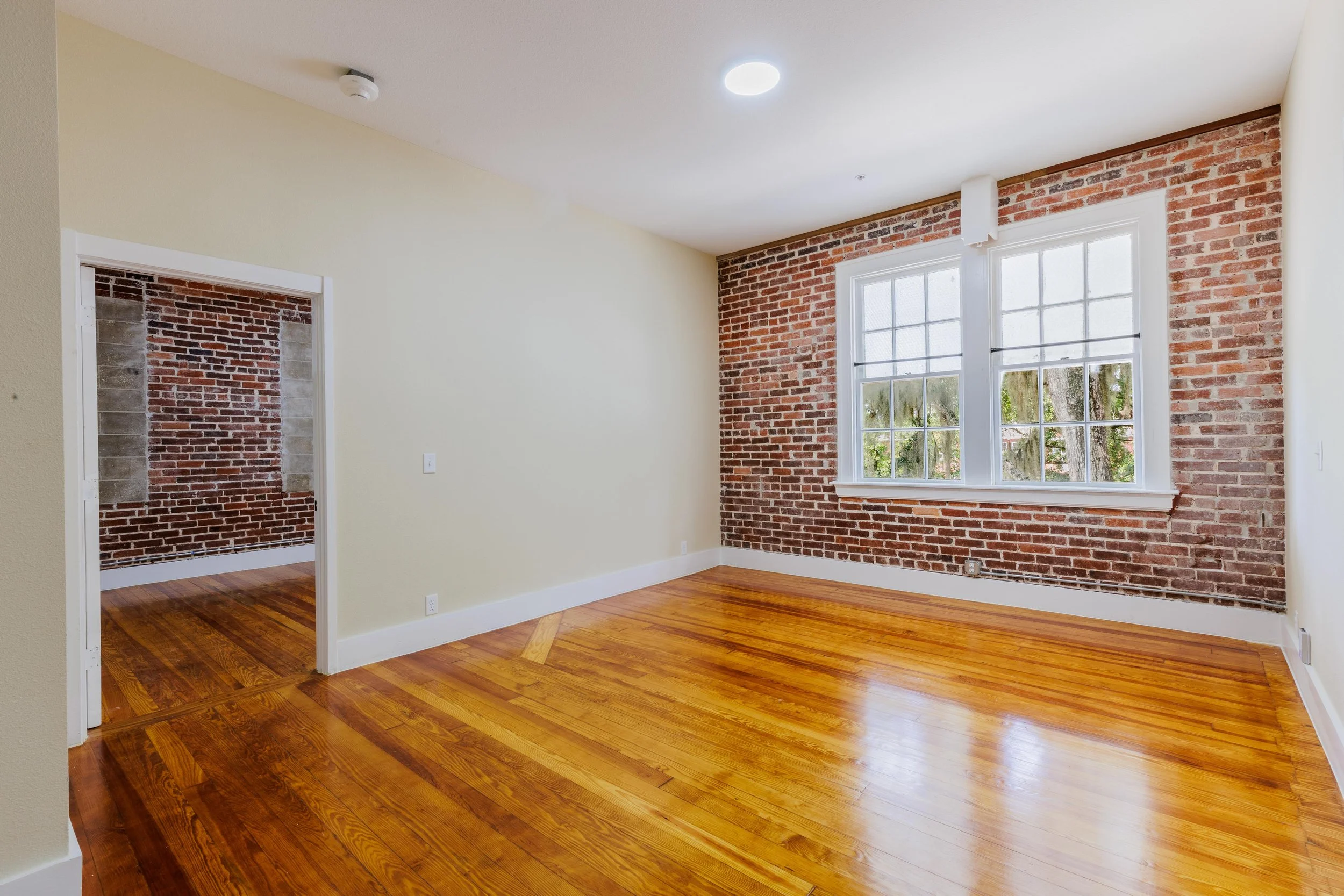 Empty room with hardwood floors, white and exposed brick walls, and large windows showing trees outside.