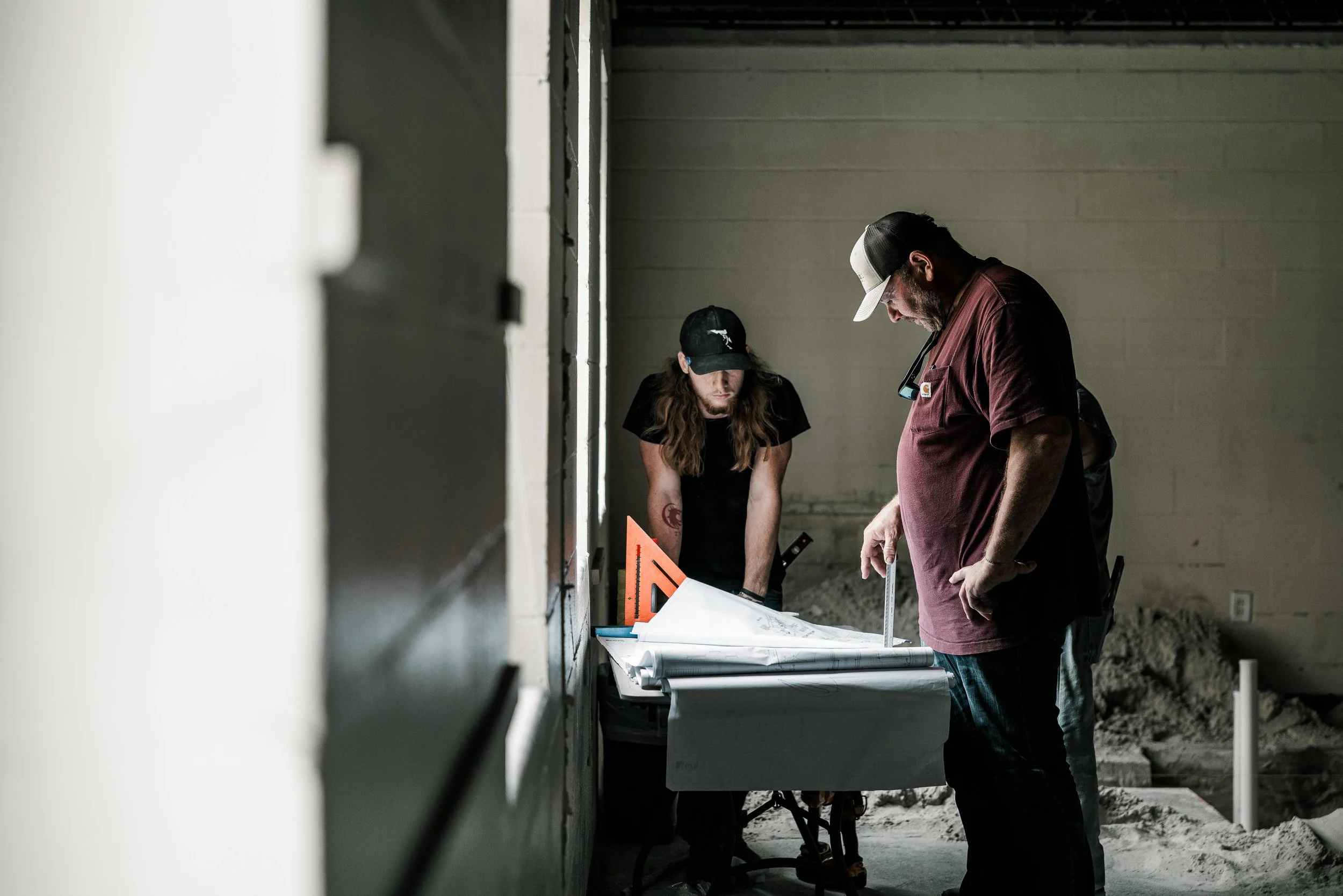Two men working on blueprints in an unfinished room with construction materials and dust on the floor.