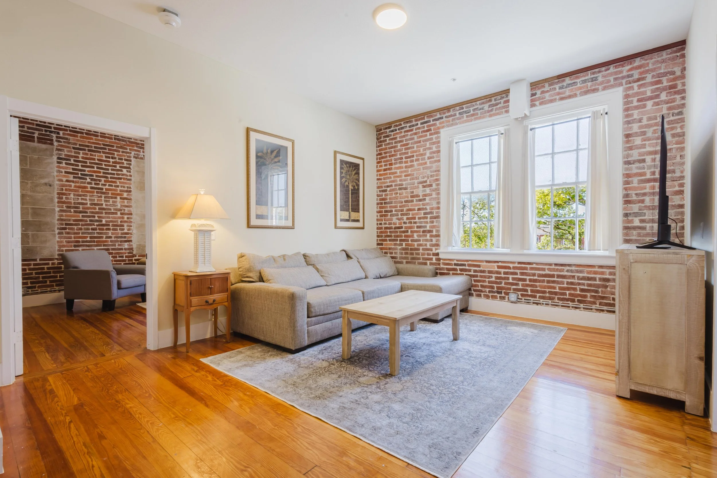 Living room with a gray sofa, wooden coffee table, cream walls, brick accent wall, large windows with white curtains, wooden floor, nearby television, and a doorway leading to an adjoining room with exposed brick wall.