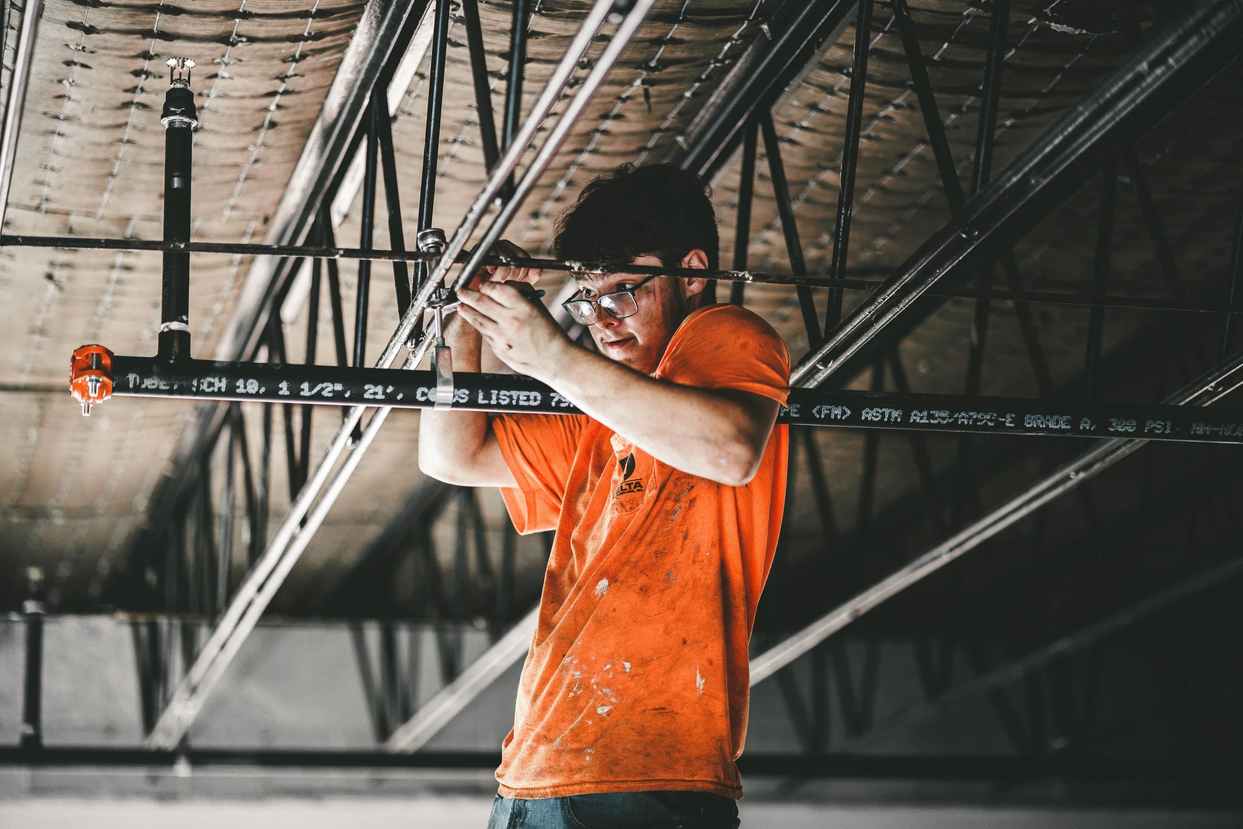 A person wearing glasses and an orange t-shirt is working on an industrial ceiling structure, using tools to assemble or repair metal framework.