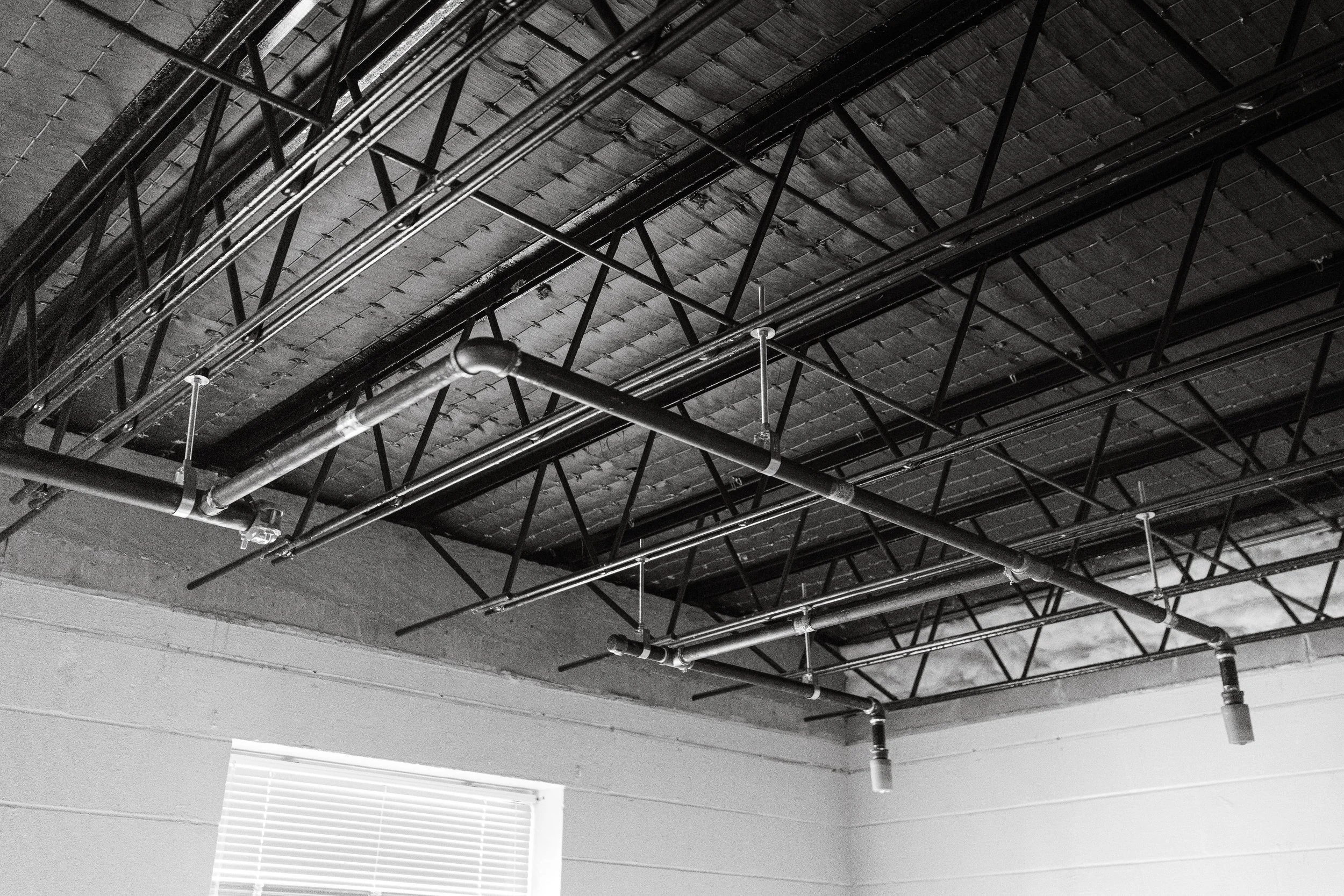 Black and white photo of an unfinished ceiling with exposed pipes and metal framework, and a window with blinds.