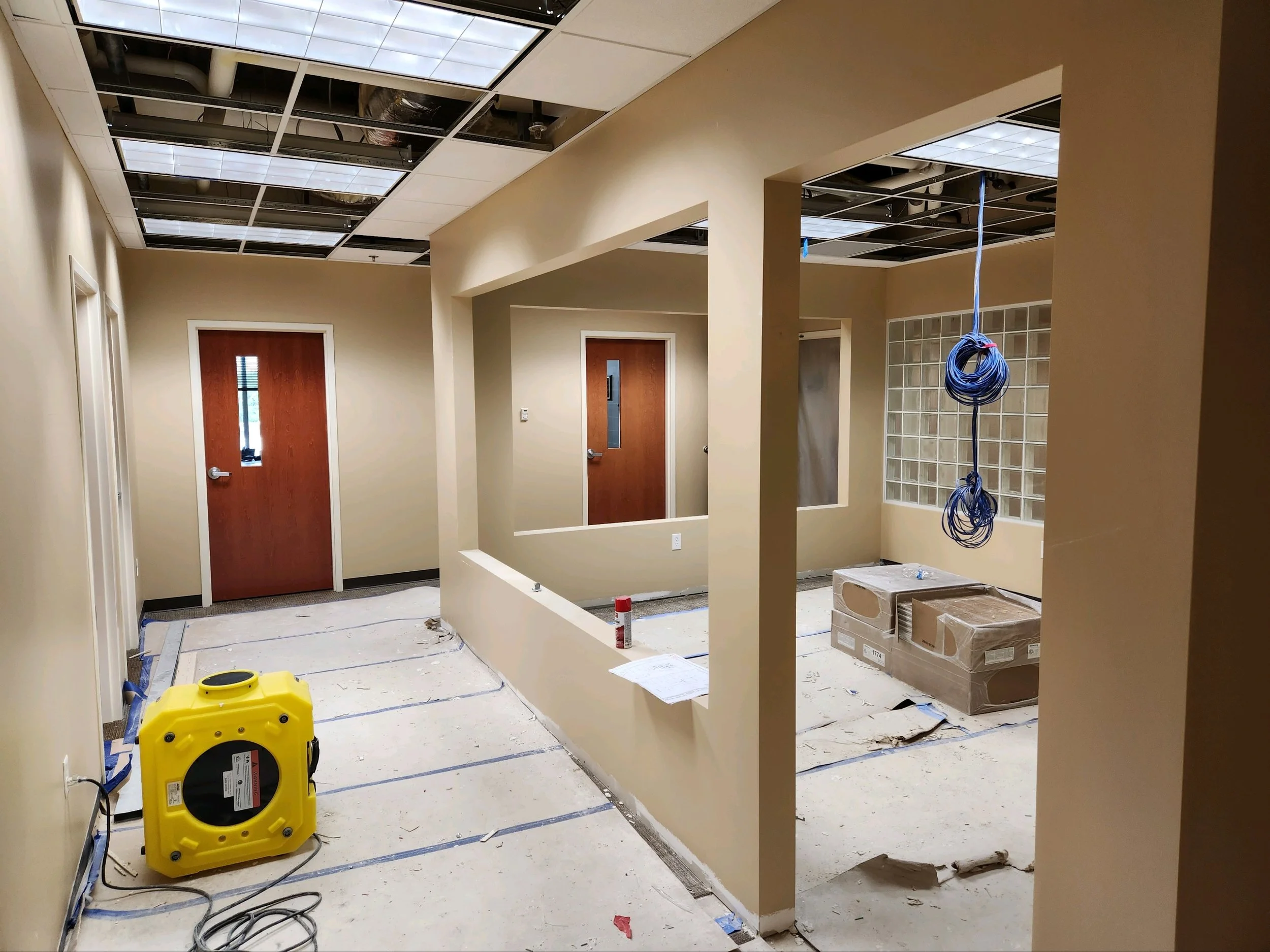 Interior of a building under construction or renovation with partially removed ceiling tiles, beige walls, and unfinished flooring, including construction tools and materials like a yellow dehumidifier, boxed items, blue electrical wires, and a layer