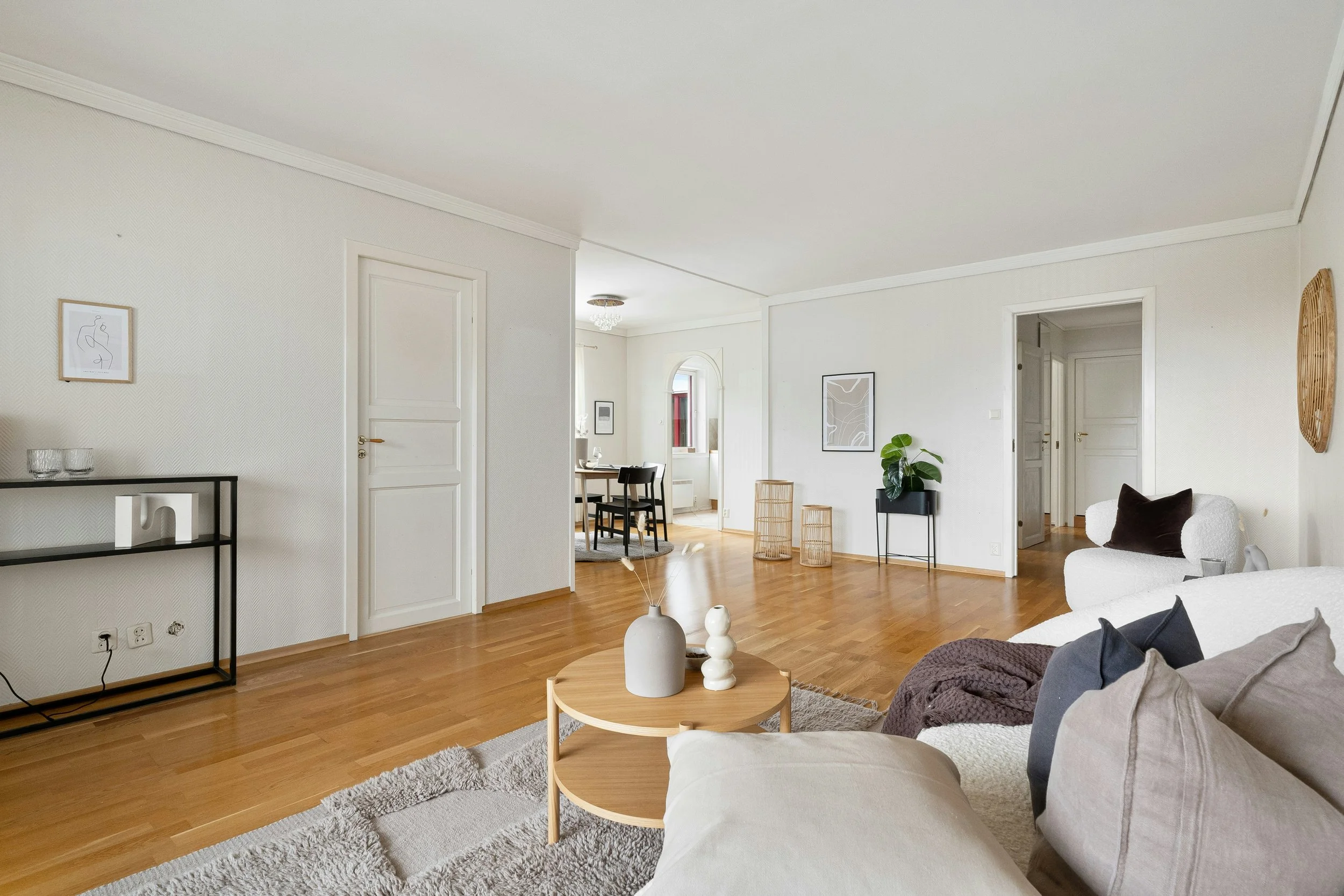 Ein hell eingerichteter Wohnraum mit weißen Wänden, Holzboden und modernen Möbeln. Es gibt einen weißen Sessel, eine kleine runde Holztisch mit Dekorvasen, sowie Wandkunst und Dekorationsgegenstände auf Regalen. Im Hintergrund sieht man einen Essbereich mit Tischen und Stühlen sowie eine kleine Pflanzen- und Dekorationsnische.