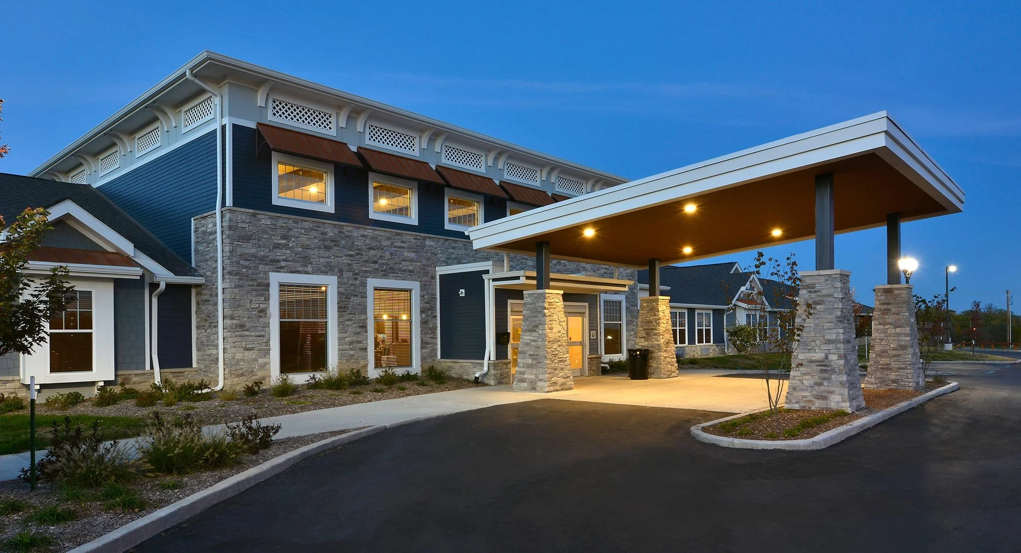 Exterior of a modern multi-story residential building with stone and blue siding, illuminated by outdoor lights at dusk, featuring a large covered entrance with stone pillars.