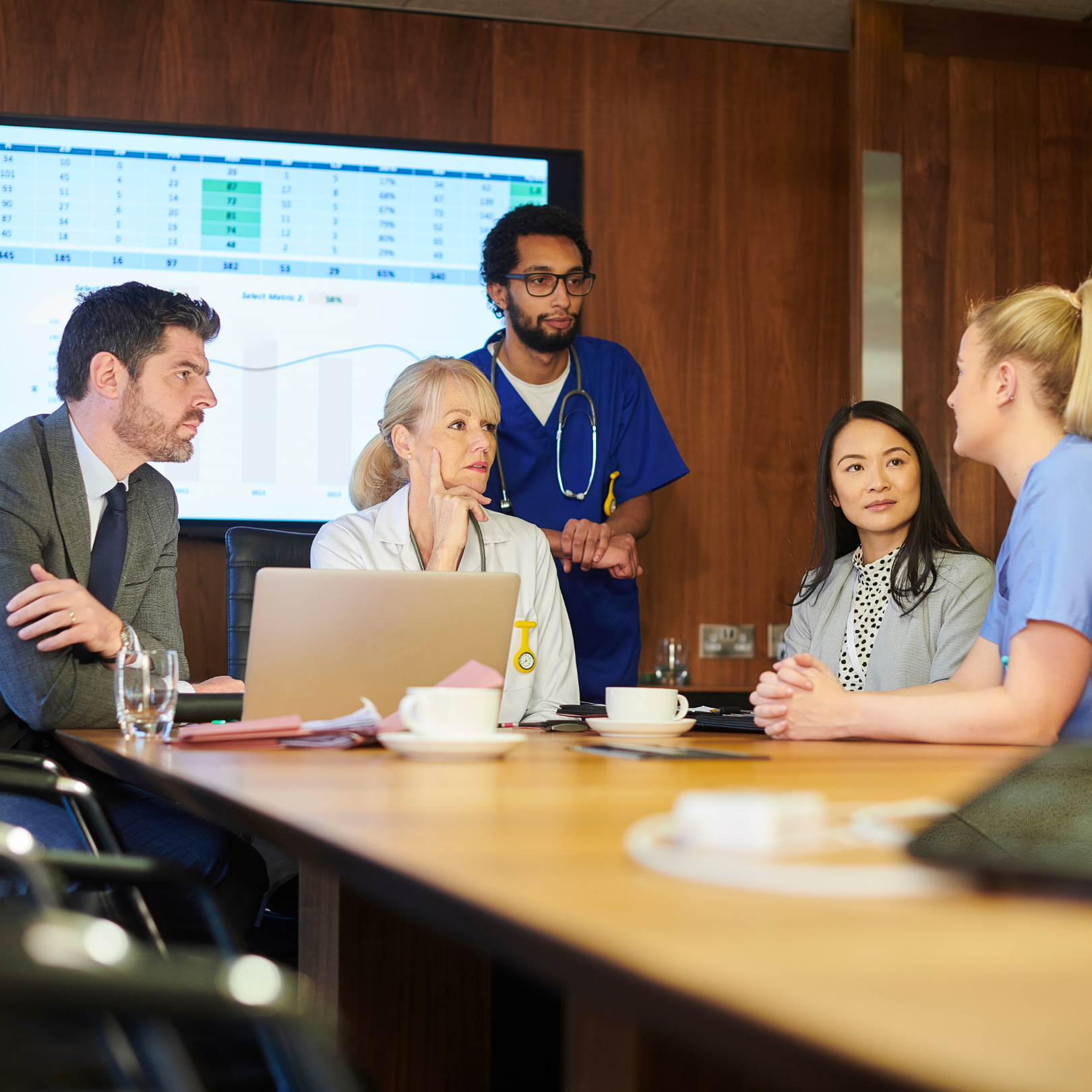 A multidisciplinary healthcare team meeting to review performance data and discuss regulatory compliance strategies in a professional conference room setting.