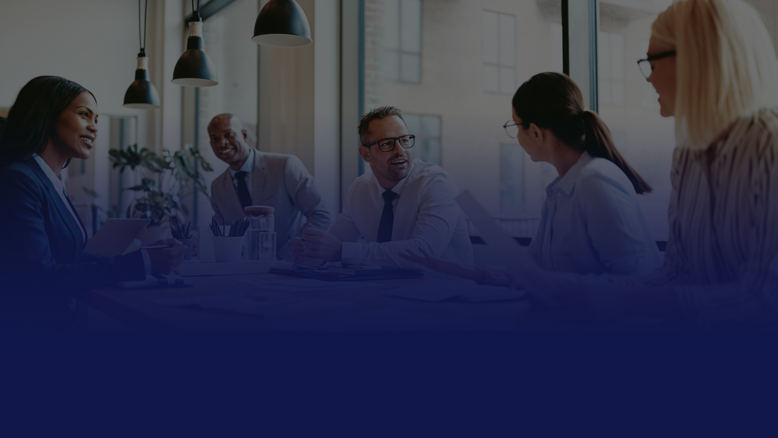 A diverse group of six professionals engaged in a meeting in an office with large windows and hanging lights.