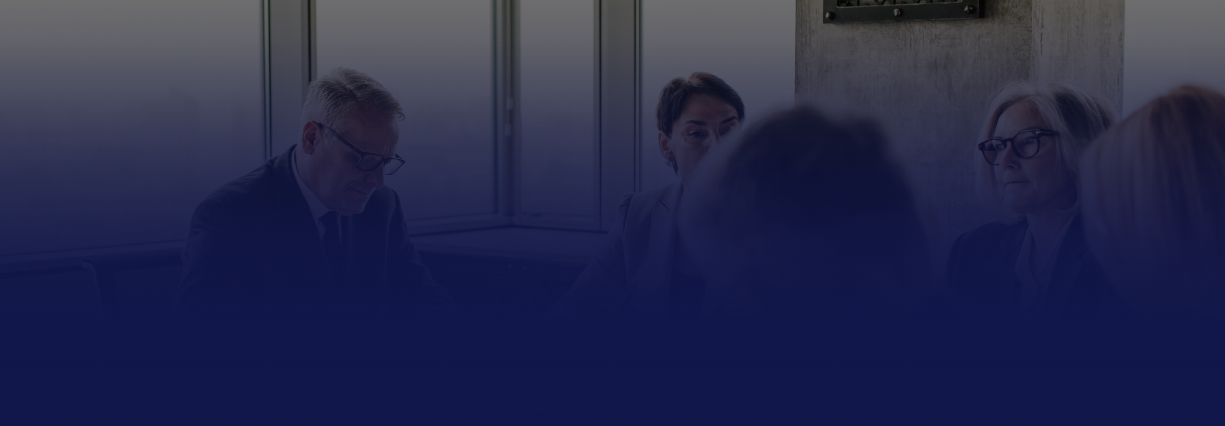 A group of five professionals in a meeting room, sitting and engaging in discussion, with a window and a wall behind them.