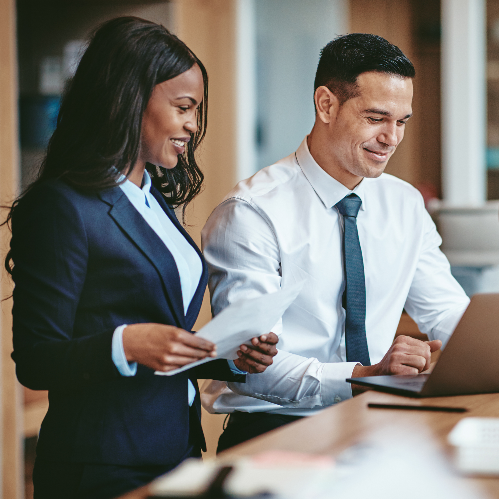 Two professionals standing side by side reviewing documents on a laptop in a modern office setting, collaborating and smiling in a professional, business-focused environment.