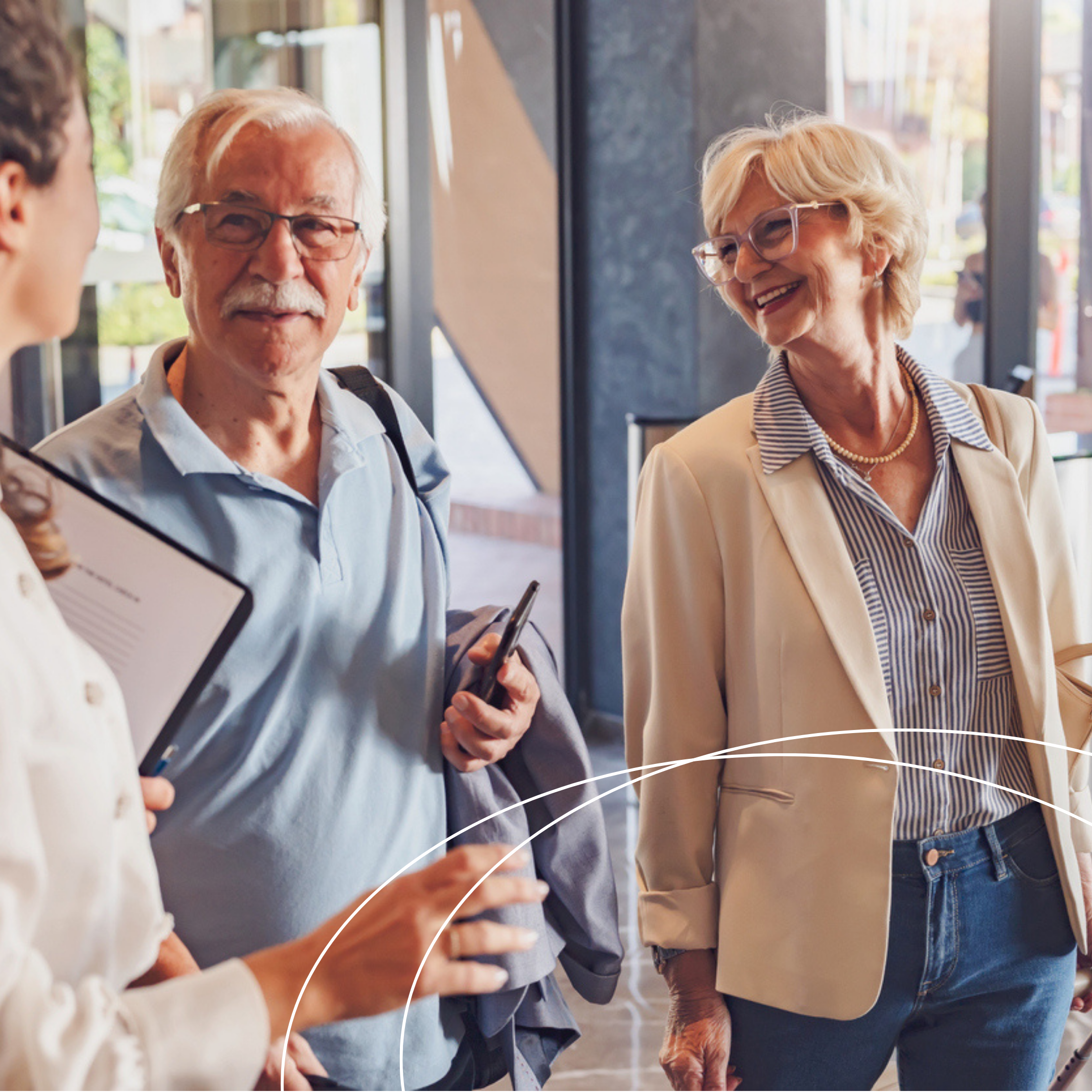 Older adults speaking with a skilled nursing accreditation professional in a lobby or office setting, holding a smartphone and tablet while engaging in a friendly conversation.