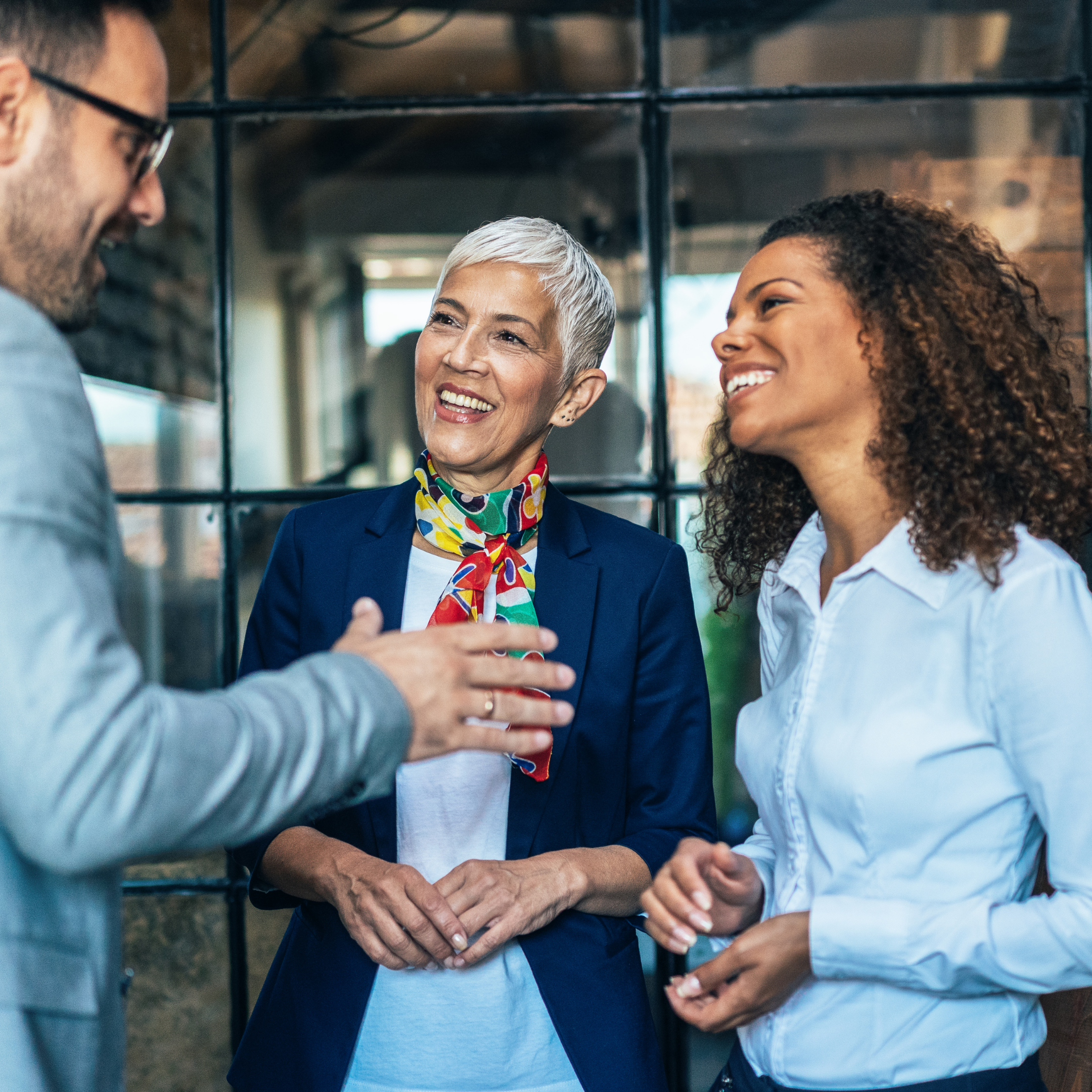 Three healthcare compliance consulting professionals standing and smiling during a conversation in a modern office setting, engaged in a friendly discussion.
