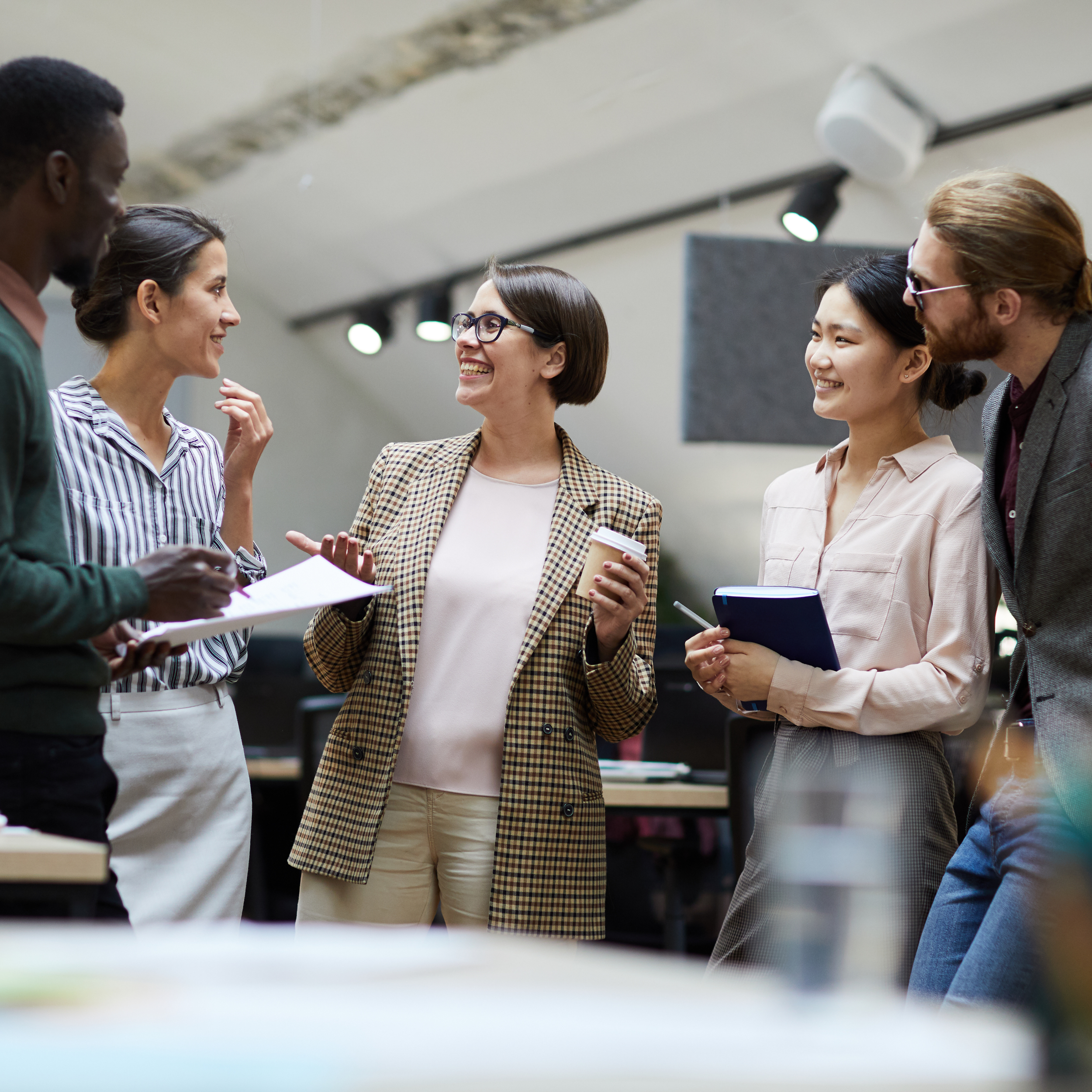Group of professionals standing together in a modern workspace, smiling and talking about accreditation support services while holding notebooks, papers, and a coffee cup.