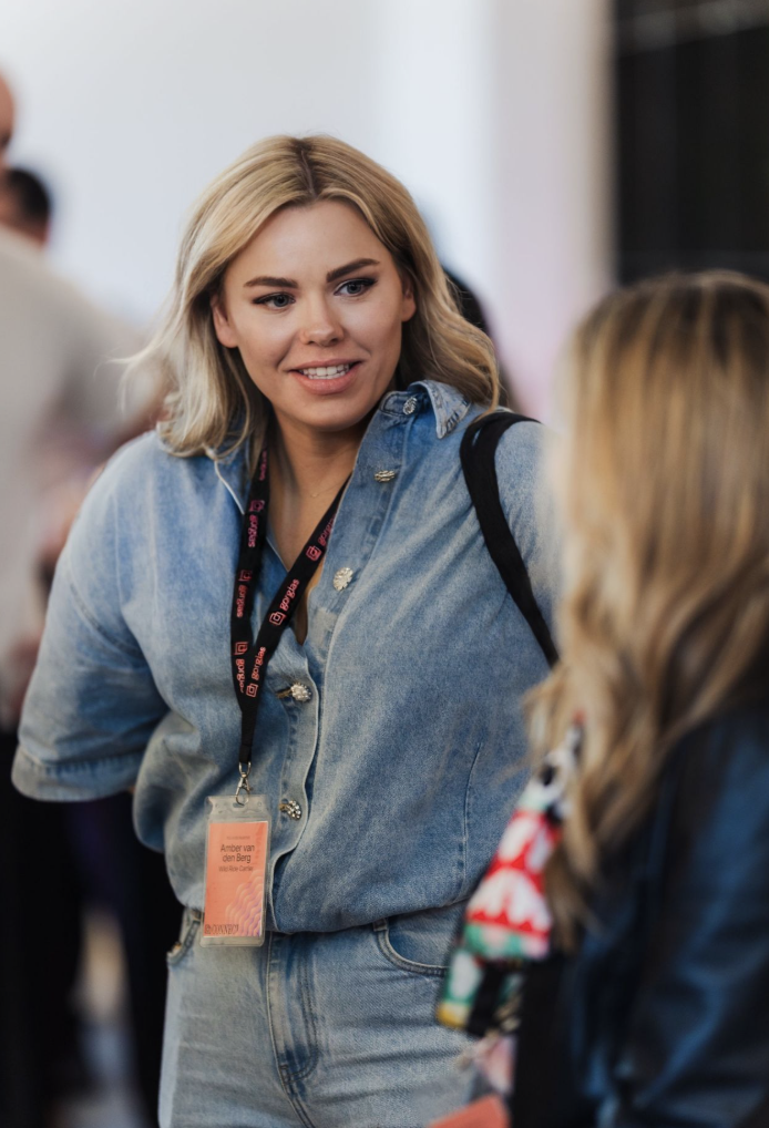 A woman with blonde hair wearing a denim jacket and a black lanyard with an orange identification badge, engaging in conversation with another person at an indoor event.