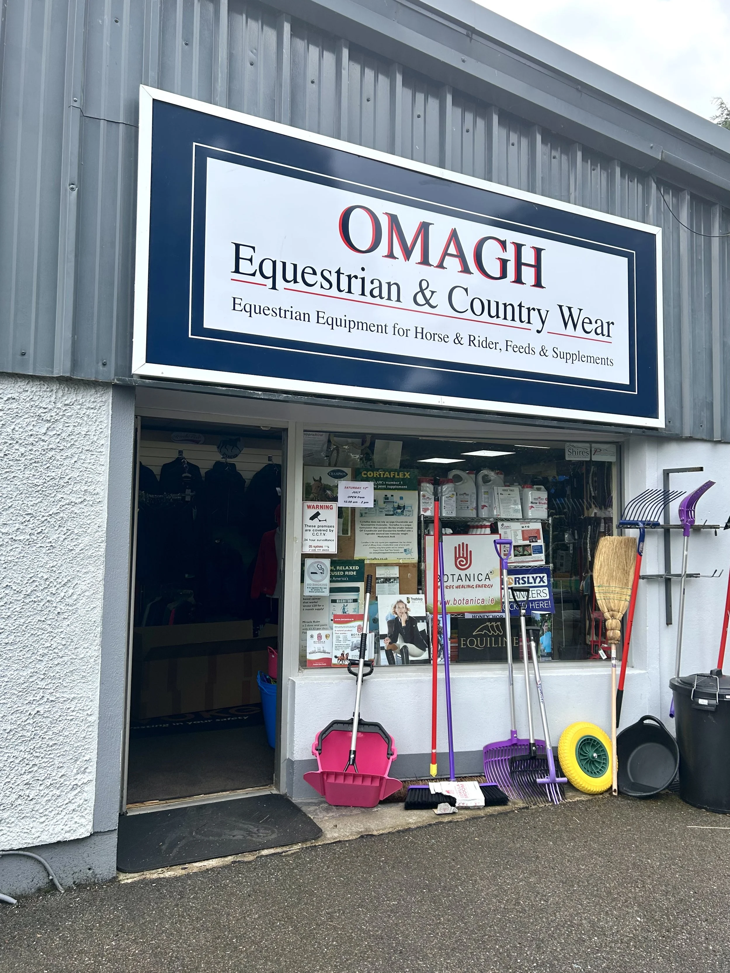 Storefront with sign reading 'OMAGH Equestrian & Country Wear' and which sells horse and rider equipment, feeds, and supplements; outdoor cleaning tools like brooms, shovels, and a wheelbarrow displayed outside.